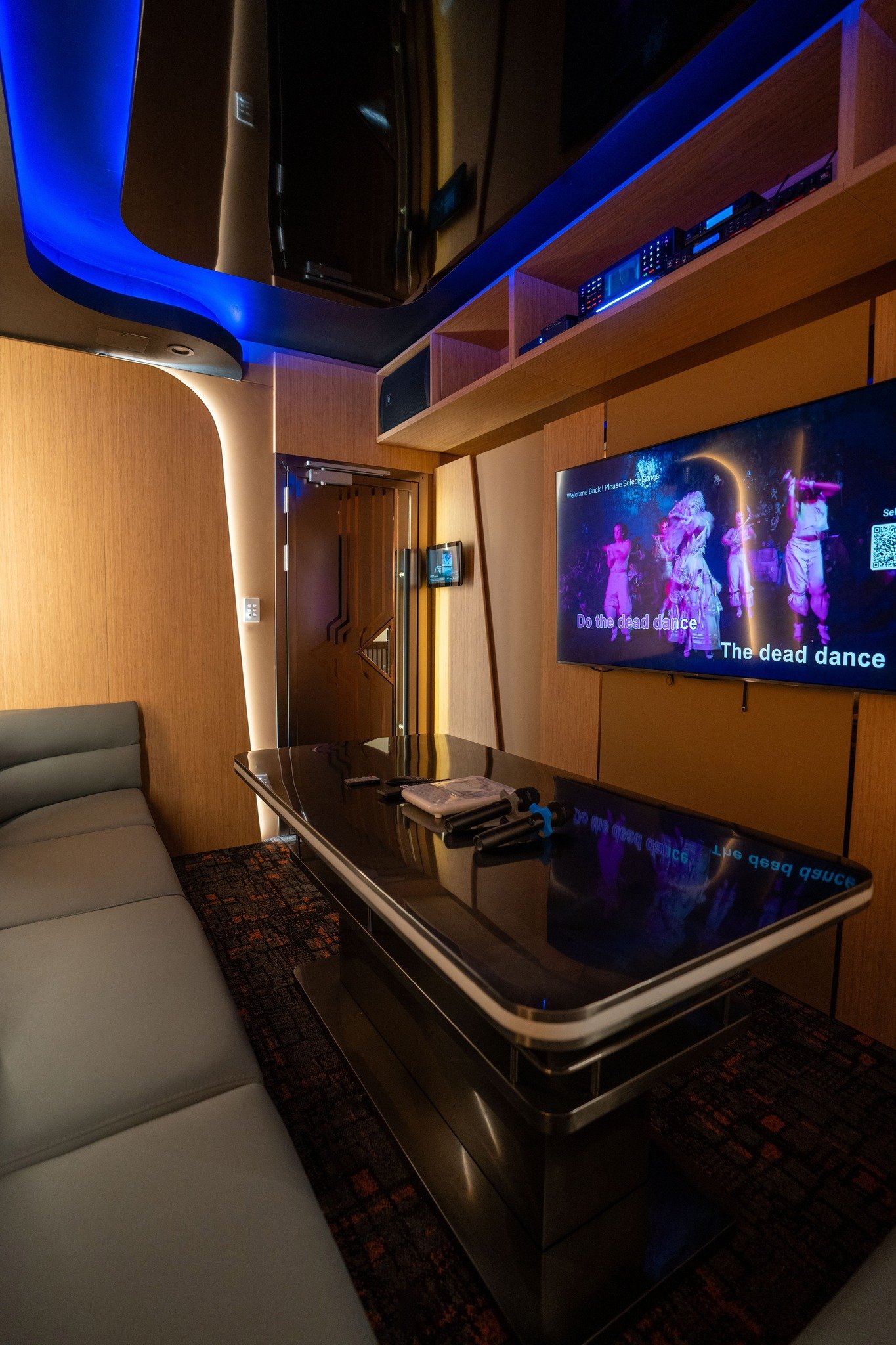 Modern lounge area with a gray sofa, a black coffee table, and a large flat-screen TV displaying a dance scene. The room features wood paneling walls, built-in wooden shelves with electronic equipment, and LED lighting accents.