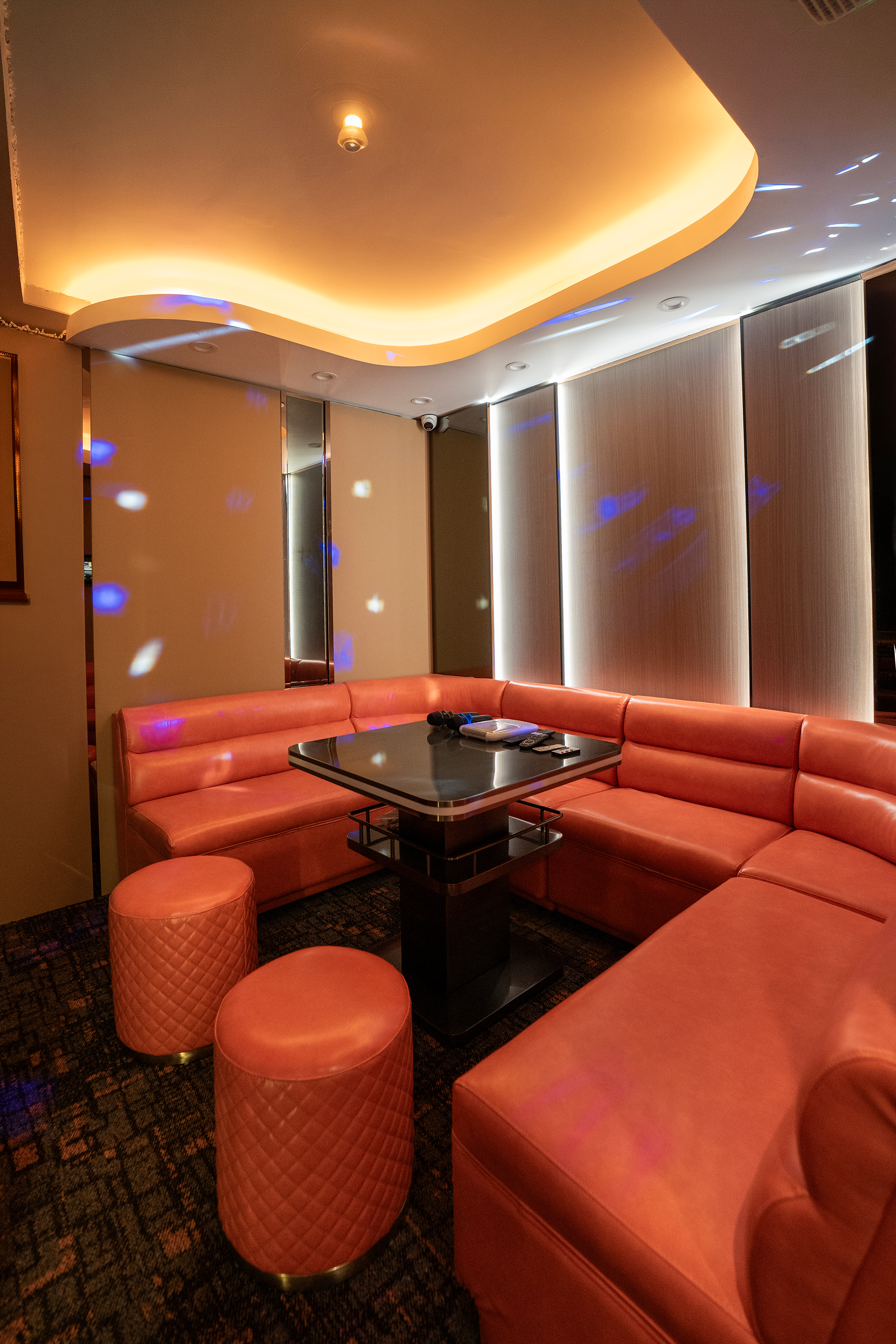 Interior of a lounge or restaurant with pink cushioned seating arranged in an L shape around a black table. Small pink quilted stools are placed nearby. The ceiling has a stylish curved recessed lighting feature. Disco lights cast blue and white reflections on the walls and ceiling. The walls are covered with beige panels, and there is a mirror on one side.