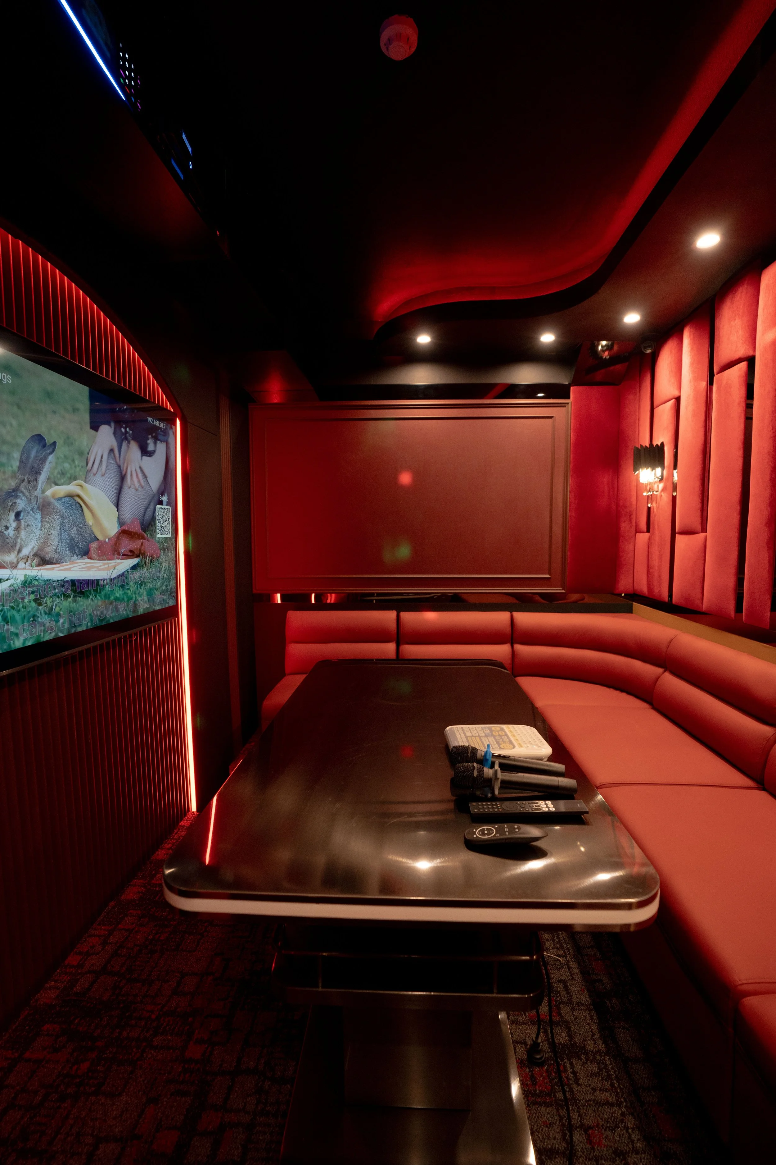 Red upholstered seating area with a large black table, remote controls, and a small gaming console. A television screen displays a rabbit and a person with a yellow scarf. The room has dim lighting with spotlights and red wall panels.