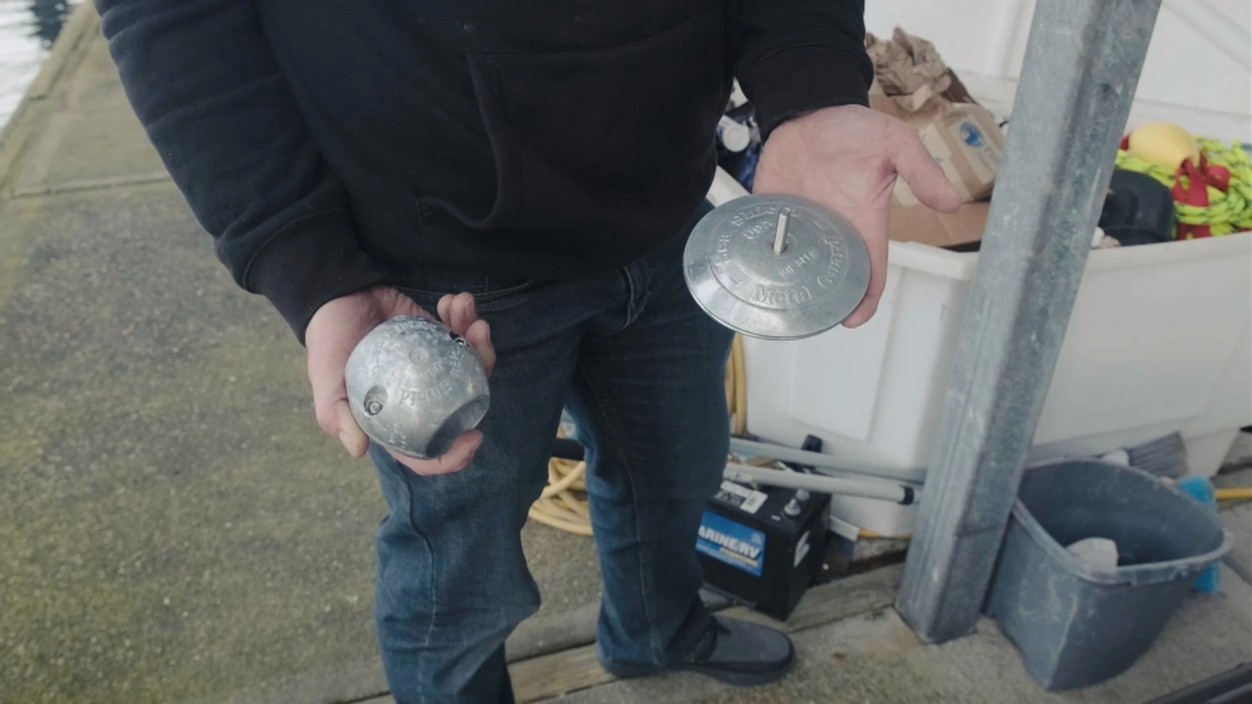 Person holding two metal weights, one in each hand, standing outdoors near a work area with tools and supplies.