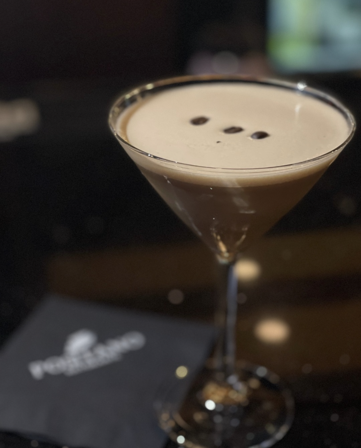 A cocktail glass filled with a creamy, brown coffee-flavored drink, garnished with three coffee beans on top, placed on a dark surface with a black napkin nearby.