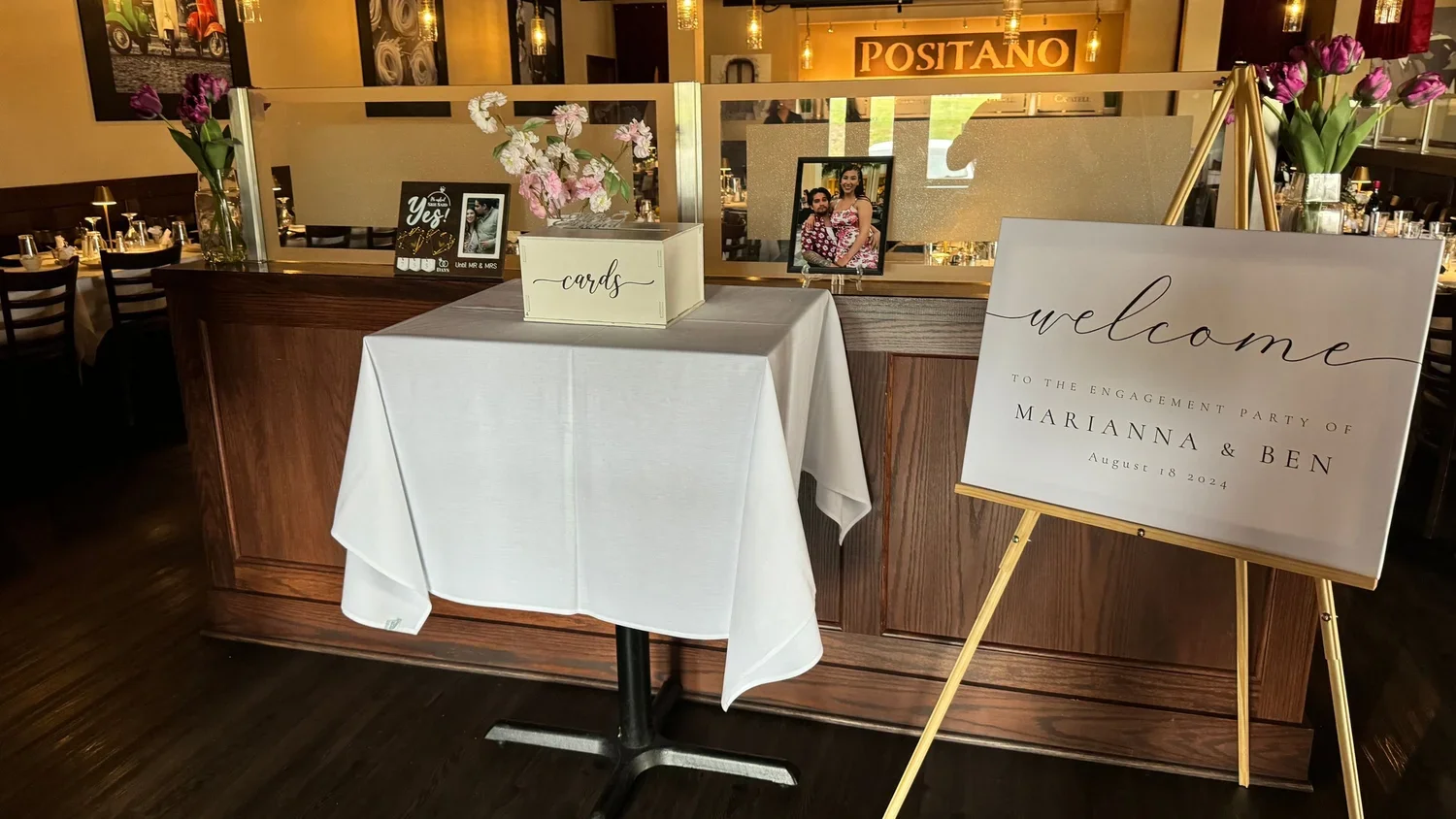 Wedding rehearsal dinner area with a welcome sign for Marianna and Ben on August 18, 2024, a table with cards and pink flowers, and a mirror reflecting a happy couple.