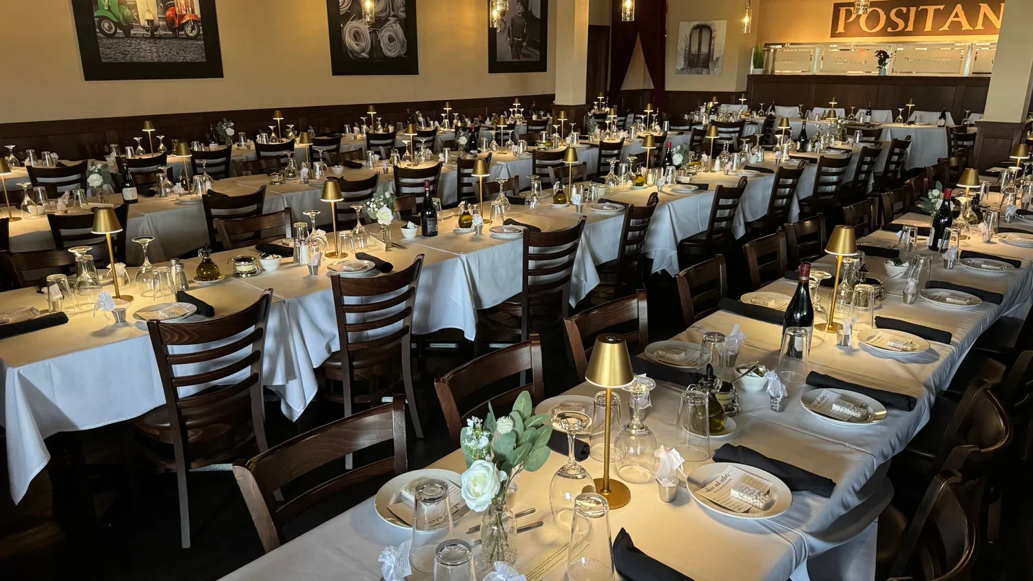 Elegant restaurant decorated with white tablecloths, gold lamps, wine bottles, glasses, and floral centerpieces, set up for a large event or banquet.