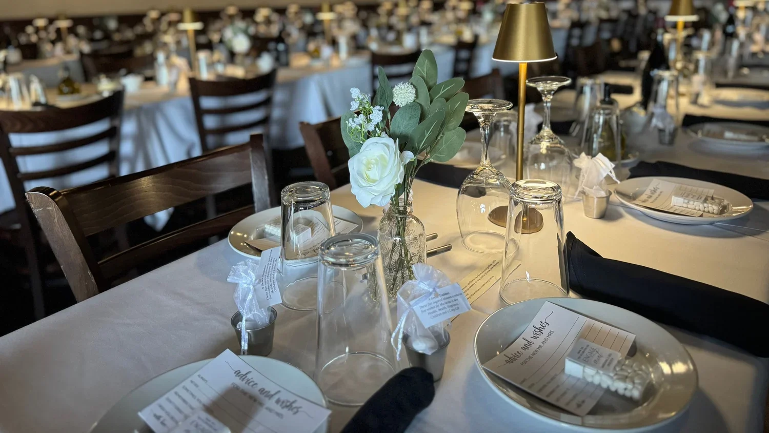 Wedding rehearsal dinner table with white tablecloth, place cards, glasses, folded black napkins, and a floral centerpiece with white roses and greenery.