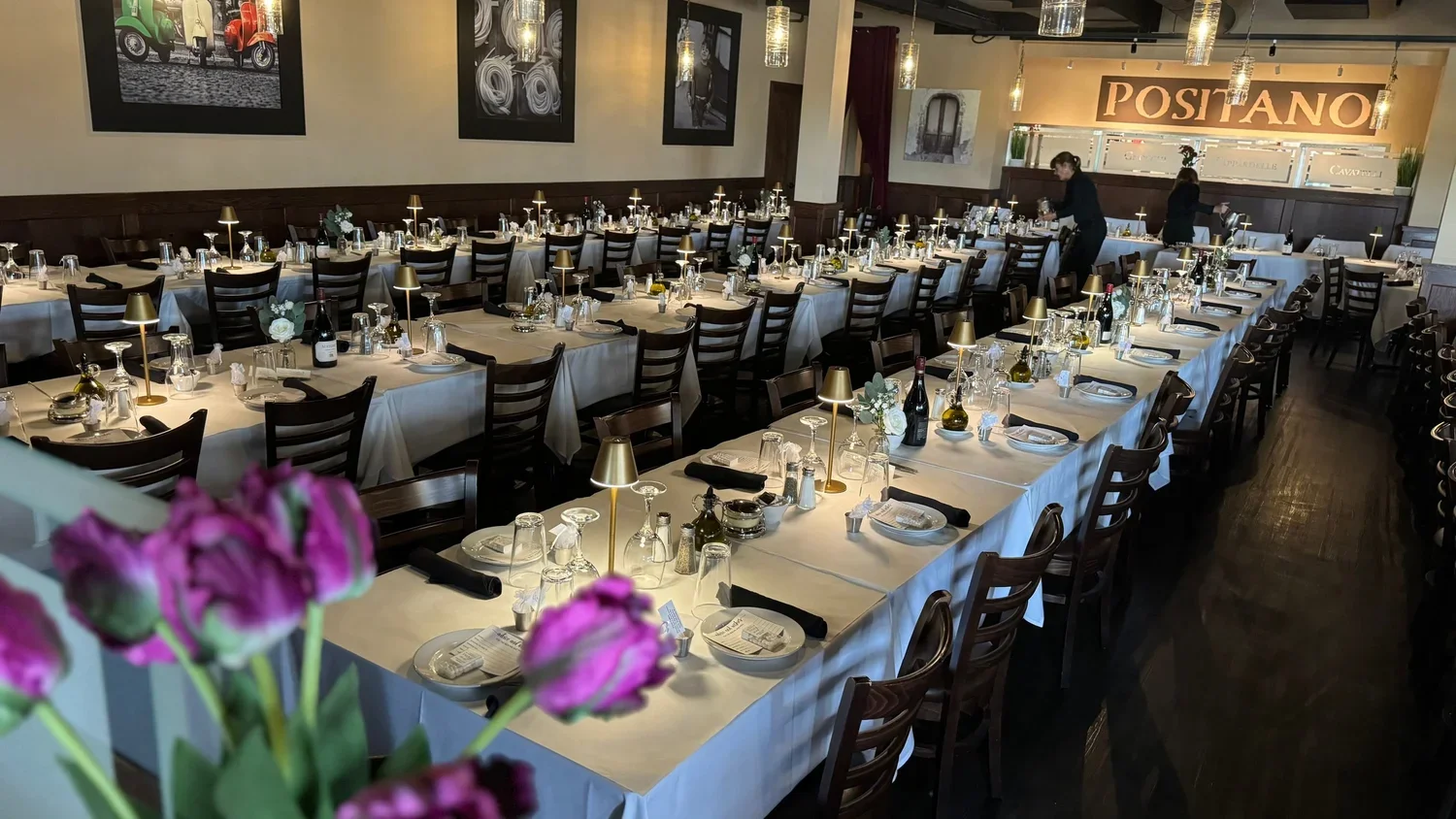 Elegant restaurant dining setup with long white-draped tables, wine bottles, glasses, white flowers, and gold lamps, with waitstaff preparing the tables. Artworks decorate the walls, and a sign reads 'POSTIANO' in the background.