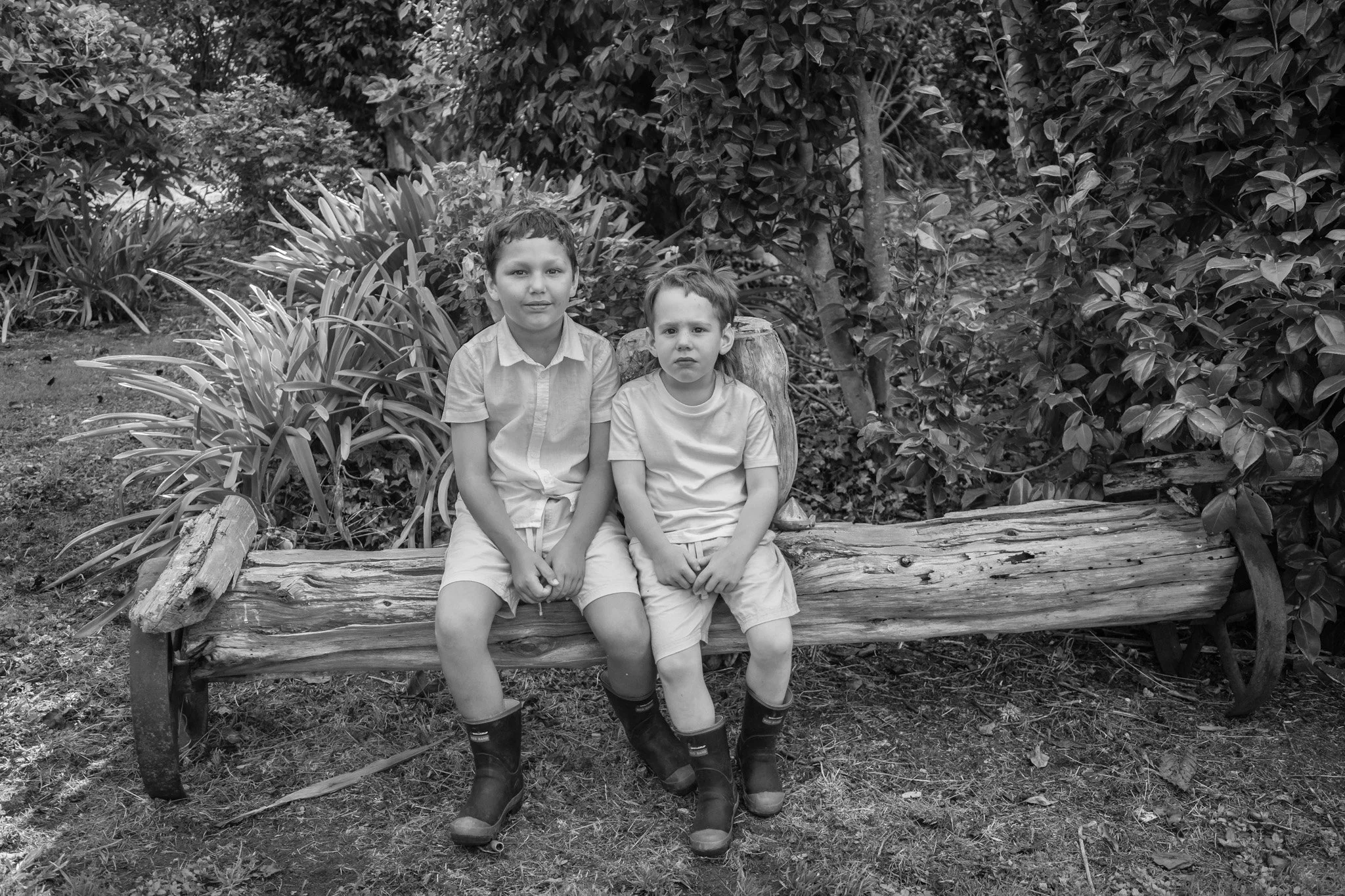 Two young boys sitting on a large log in a garden or park, surrounded by plants and trees.