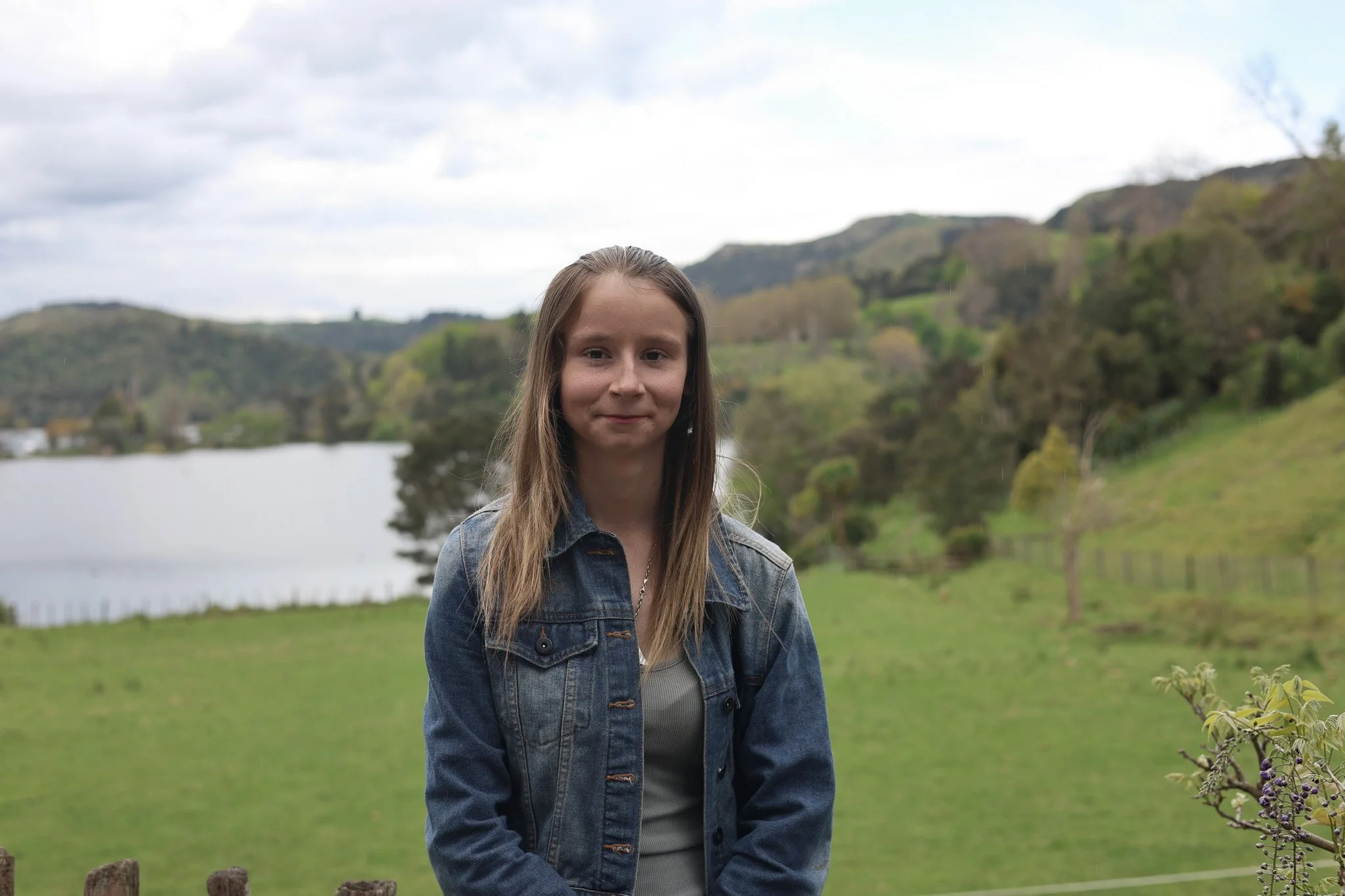 A young woman with long brown hair standing outdoors near a lake and green hills, wearing a denim jacket and a light-colored top, smiling softly.