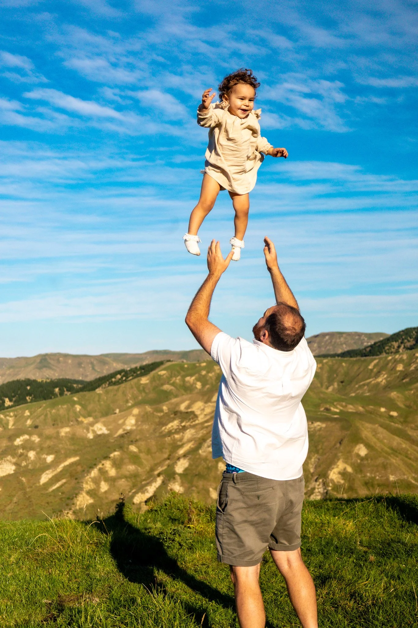 A man tossing a young girl into the air outdoors with mountains and a blue sky in the background.