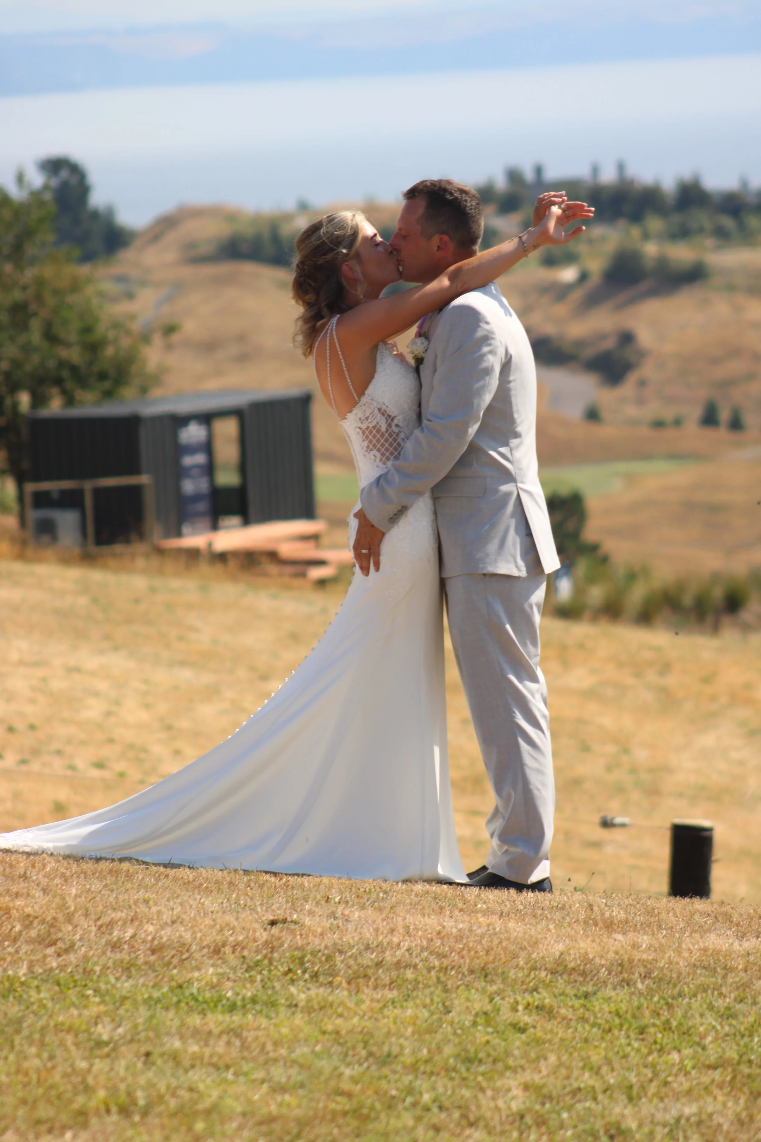 A newlywed couple sharing a kiss outdoors on a grassy hill, with rolling hills in the background.