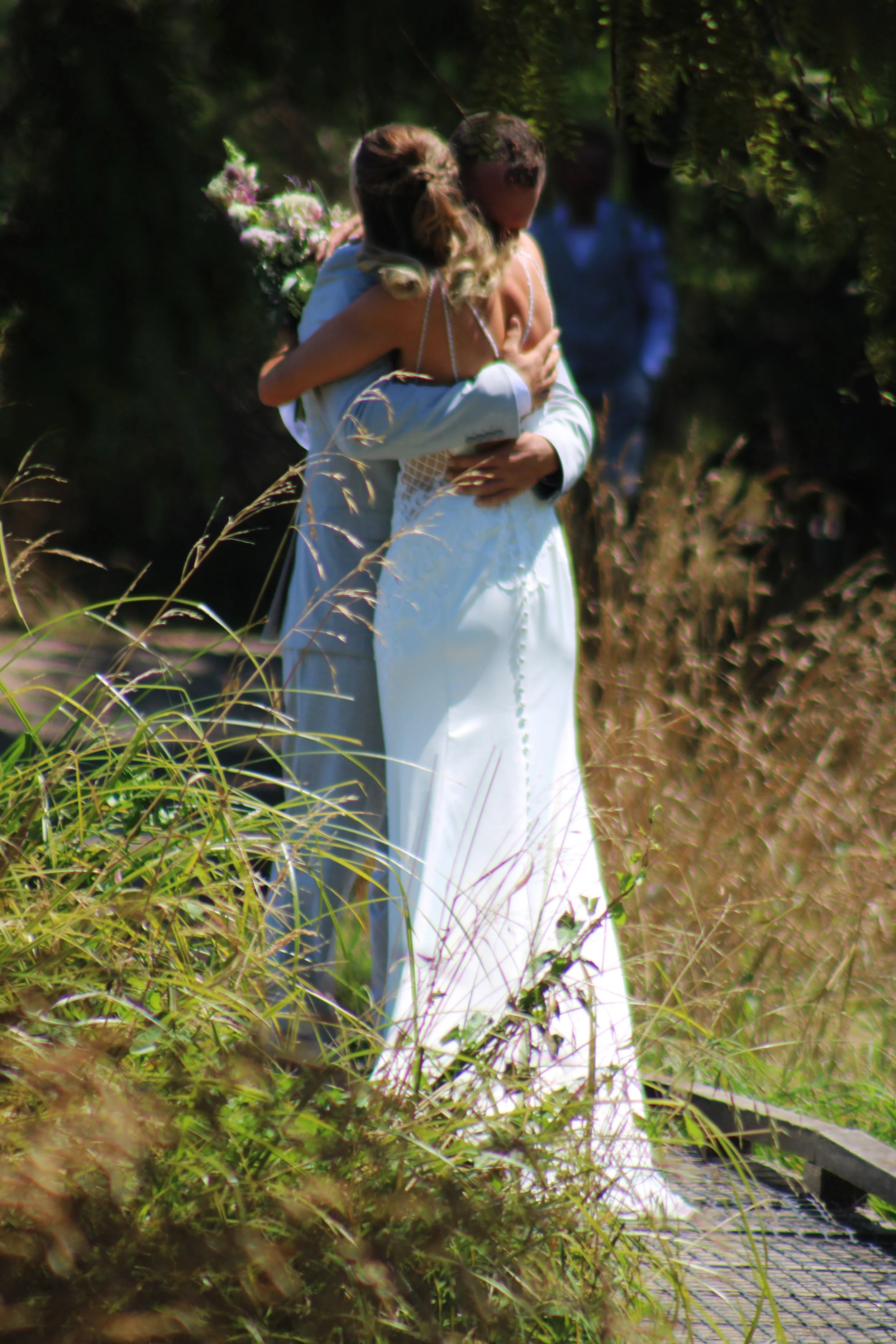 A bride in a white dress hugging a groom in formal attire outdoors on a grassy path surrounded by tall grass and trees.