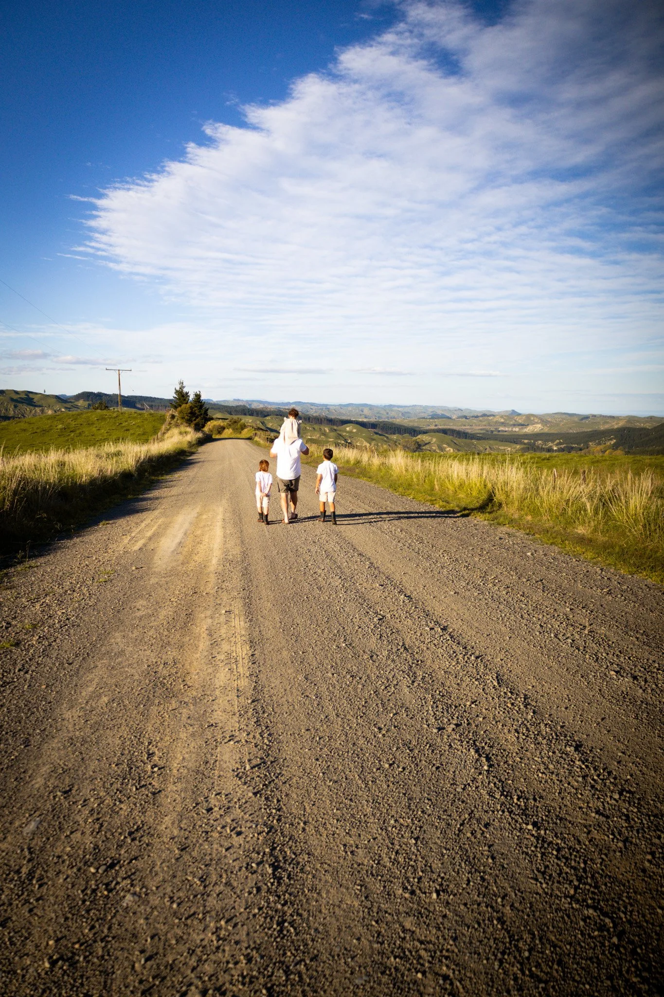 A person walking with two children on a dirt road through a grassy landscape under a partly cloudy sky.