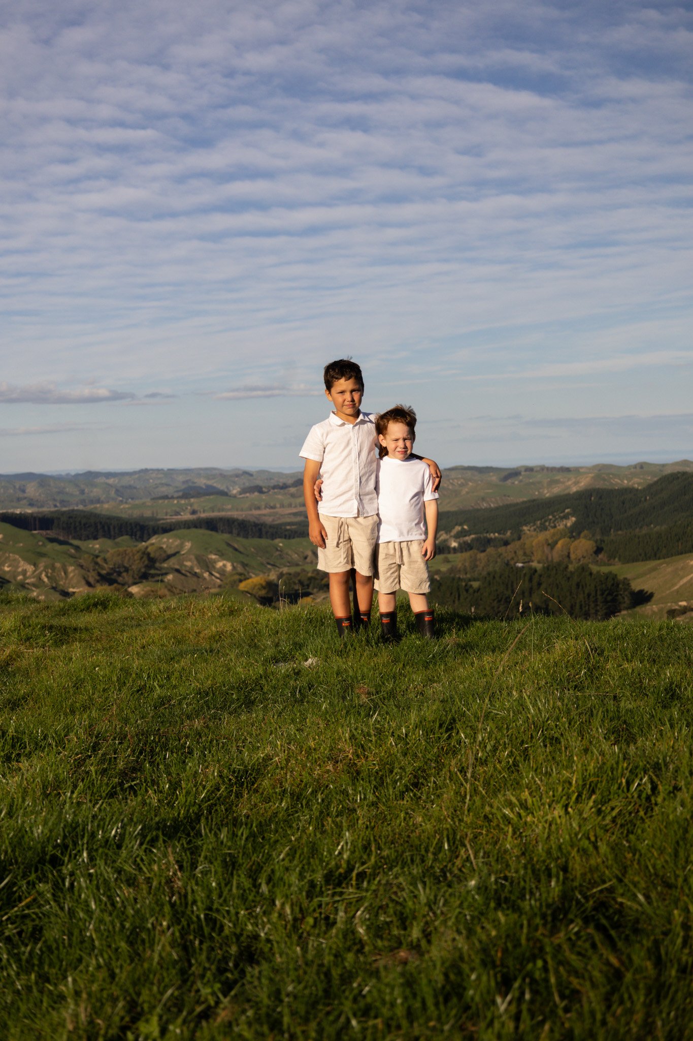 Two boys standing on a grassy hill with scenic rolling hills and a blue sky with clouds in the background.