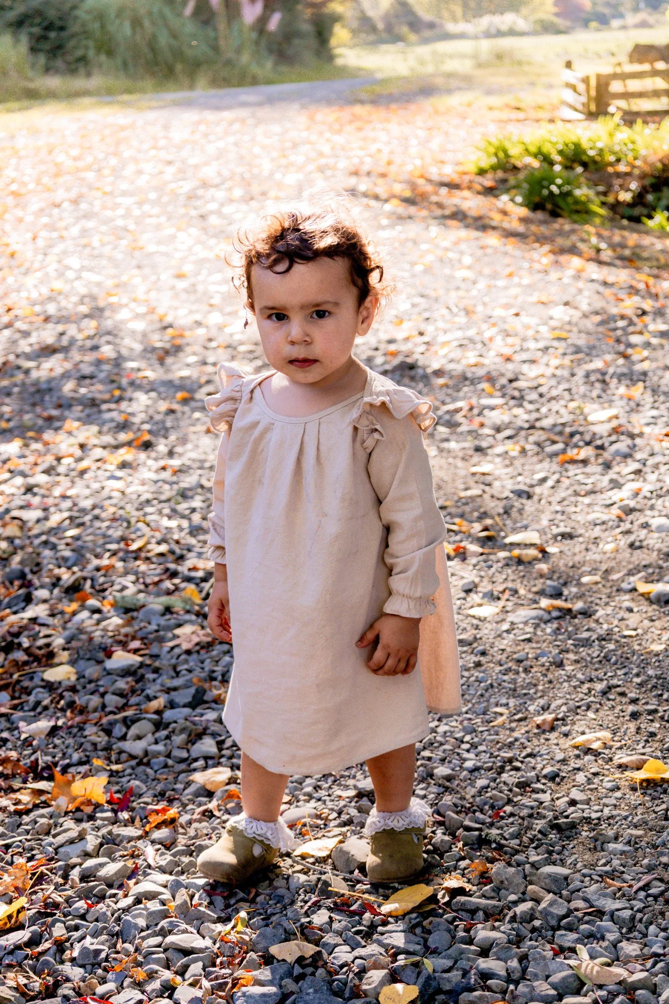 A young girl with curly hair and a serious expression, wearing a beige dress and shoes, standing on a rocky pathway outdoors during autumn.