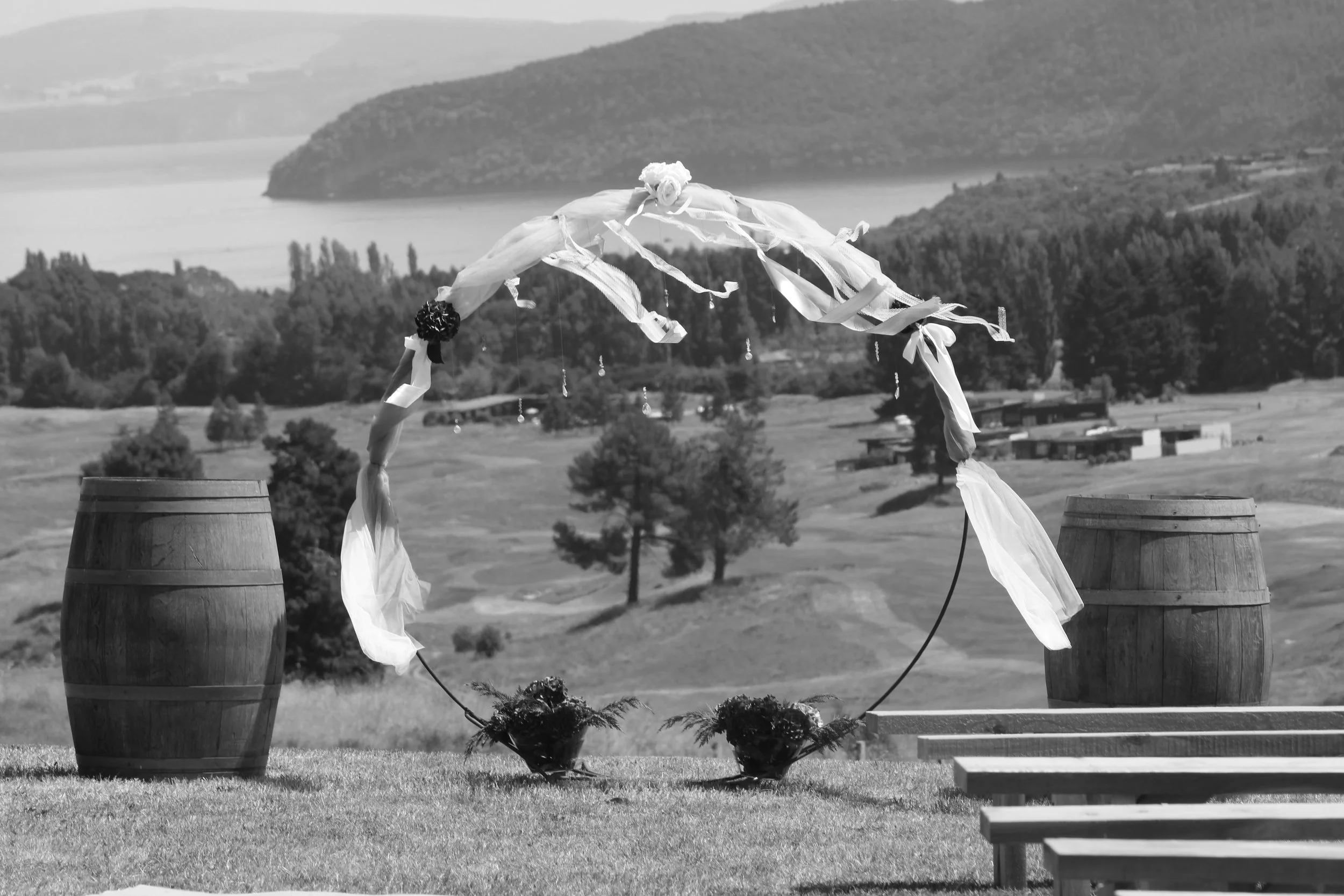 A wedding arch decorated with fabric and flowers, flanked by two wooden barrels, set on a grassy hill with a lakeside view in the background.