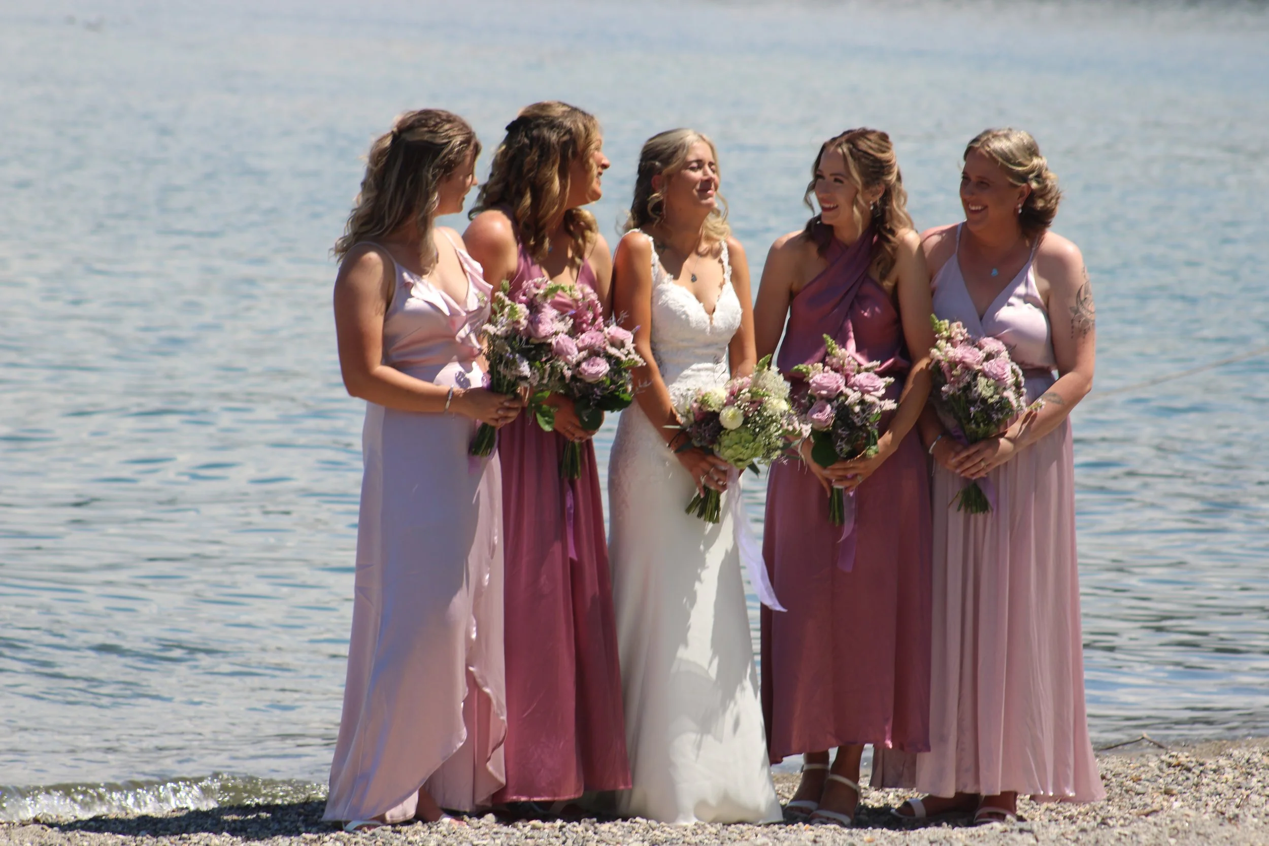 A bride in a white wedding dress standing on a beach with five bridesmaids in pink dresses, all holding bouquets of flowers, near the water.