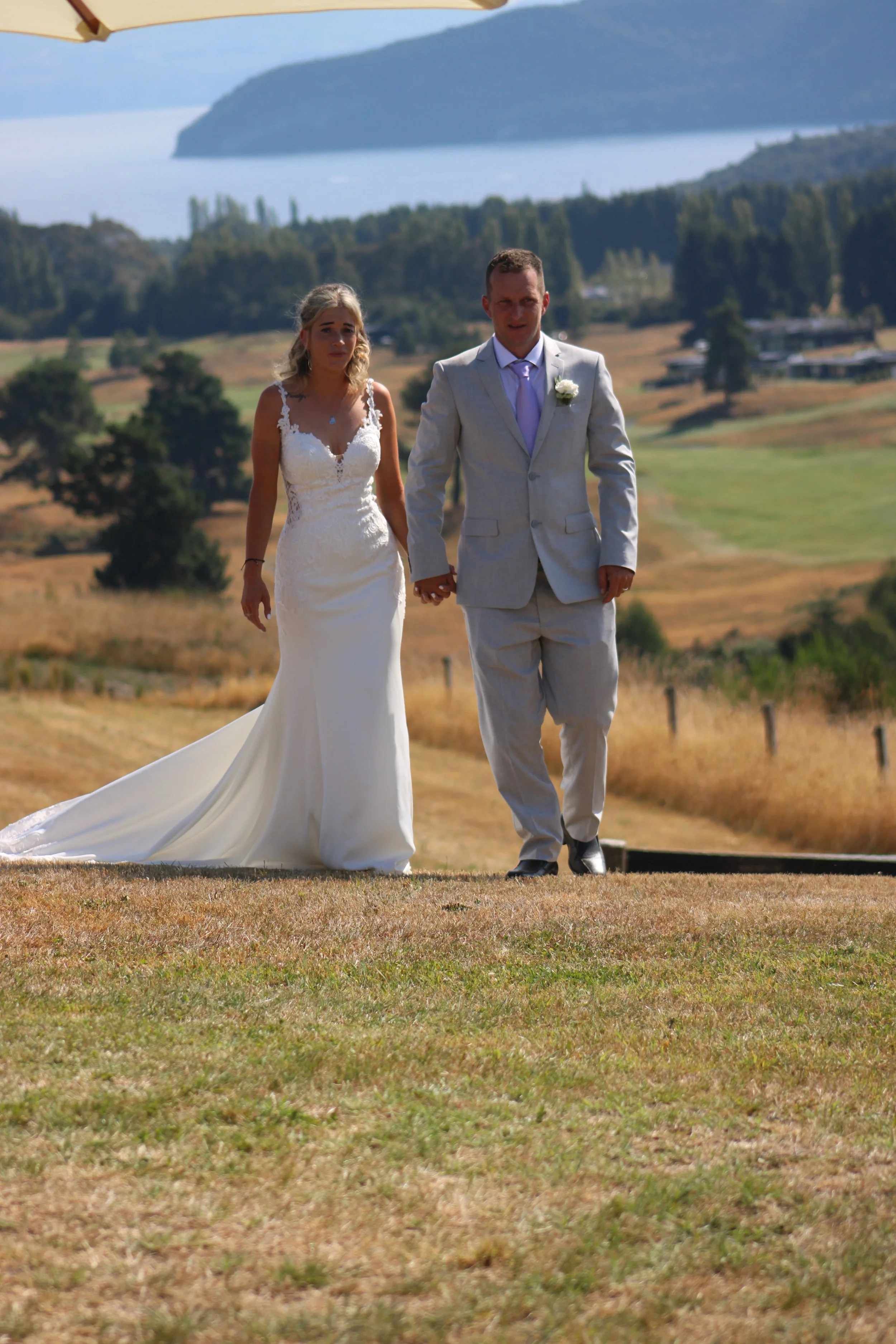 A bride and groom walking hand in hand outdoors on a sunny day, with a scenic landscape of water, hills, and trees in the background.