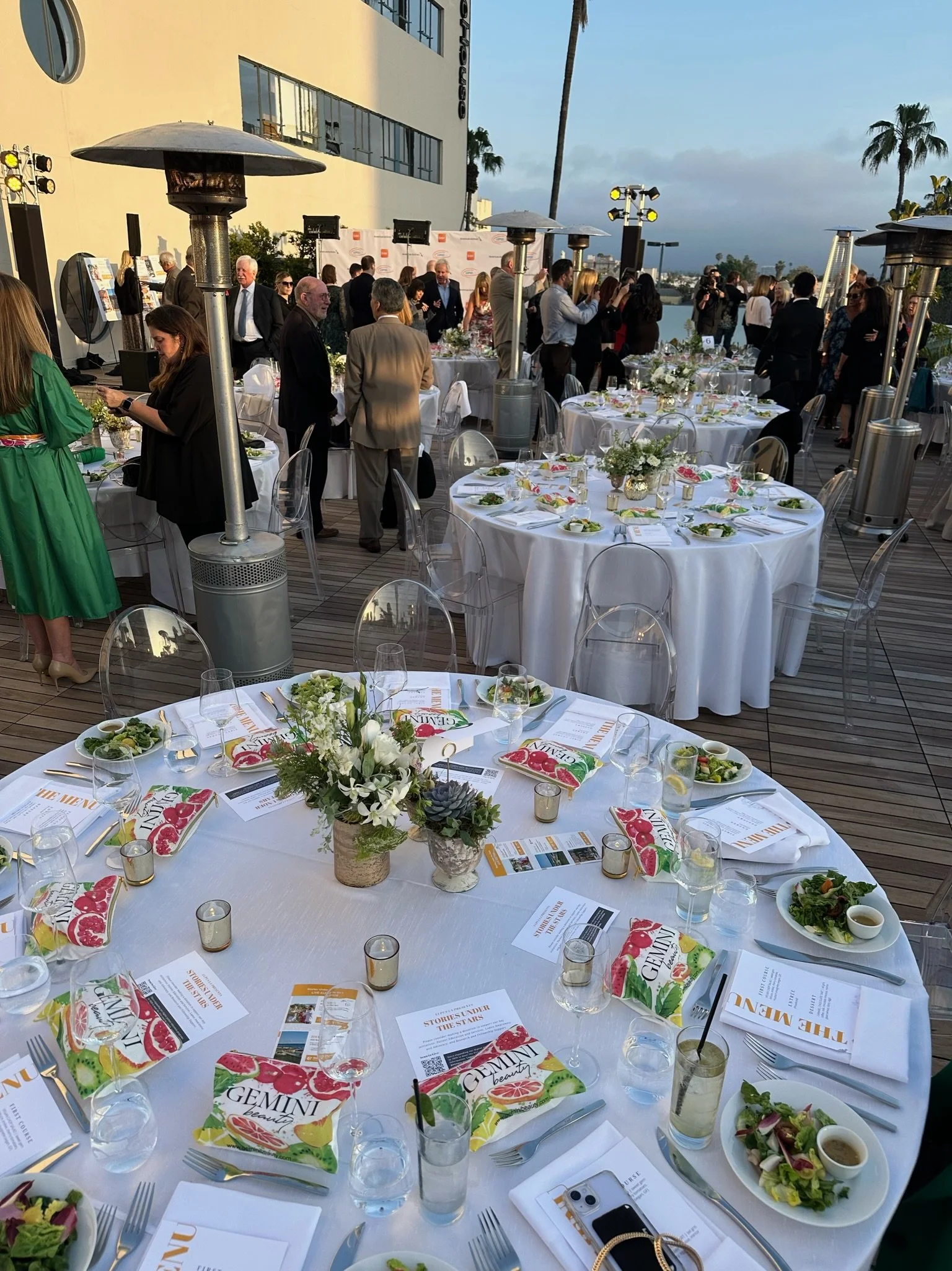 Outdoor event with round tables set with white tablecloths, floral centerpieces, salads, menus, glassware, and candles, with people mingling and tall patio heaters visible.