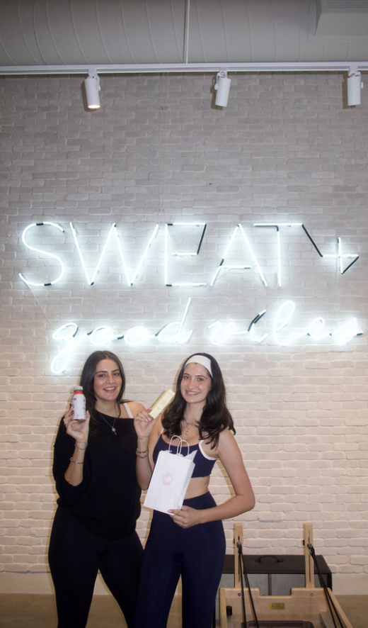 Two women smiling and holding health and wellness products, standing in front of a neon sign that reads 'SWEAT good vibes' on a brick wall.