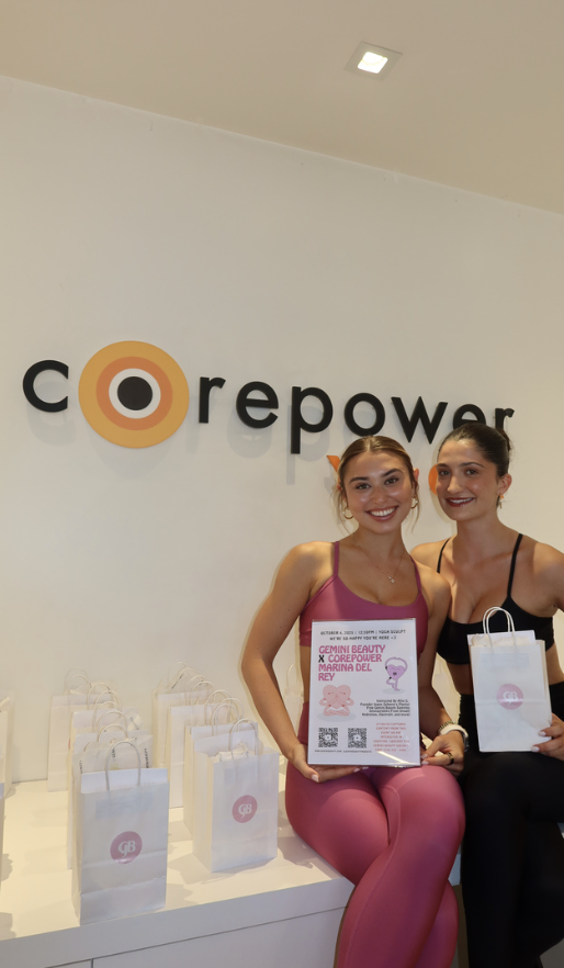 Two women smiling and sitting in front of a sign that says 'corepower,' with white gift bags on a table in front of them.