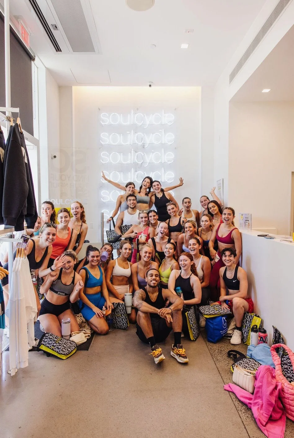 A group of fit young women and men in workout clothes gathered inside SoulCycle studio, posing for a group photo in front of a illuminated wall with 'soulcycle' repeated on it.
