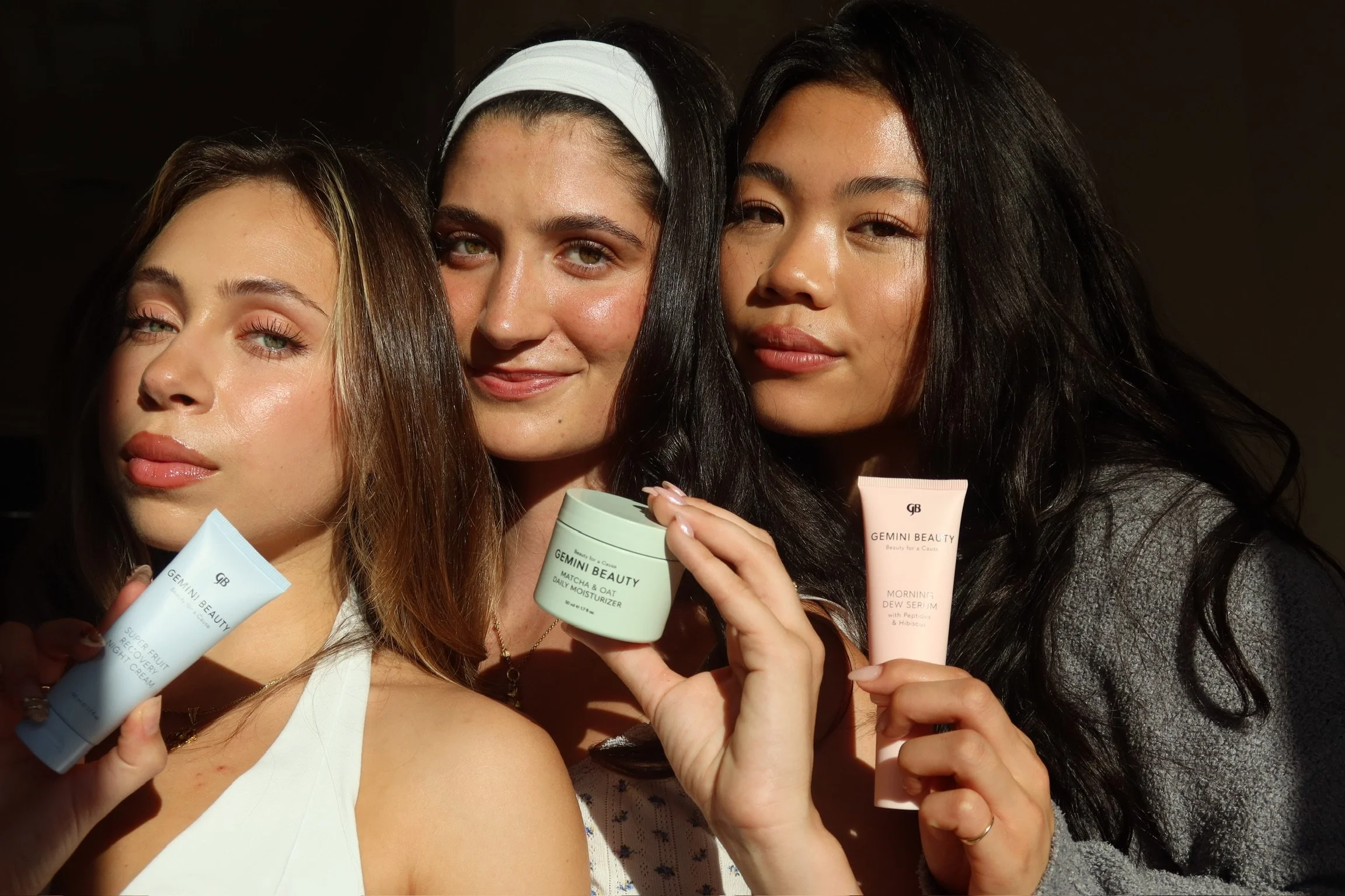 Three women holding skincare products, smiling and posing in natural light.