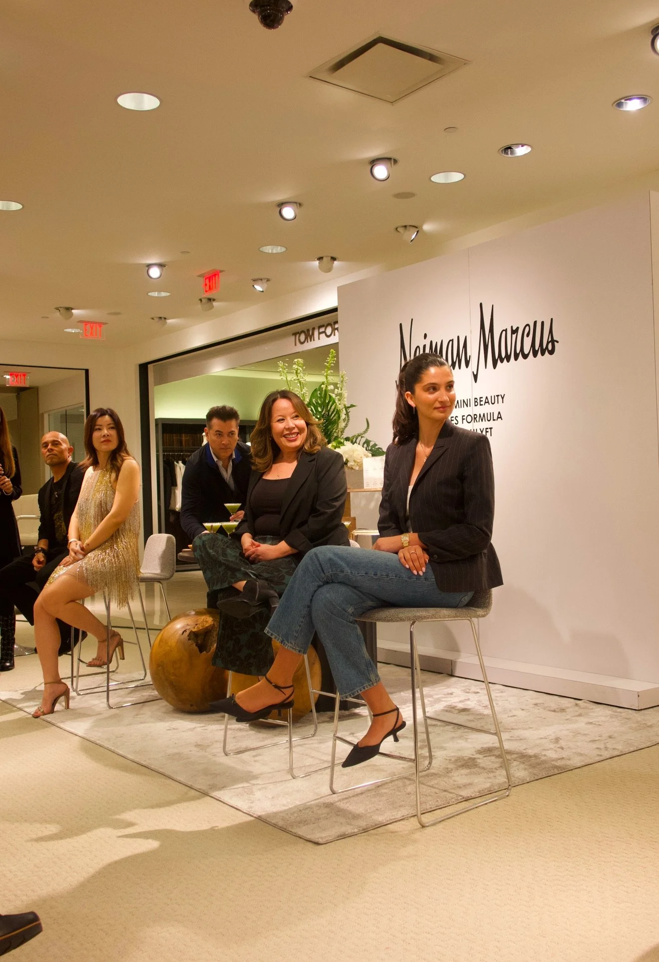 People sitting on chairs at a panel or event in a retail store, with a large sign that reads 'Naima Marcus' in the background.