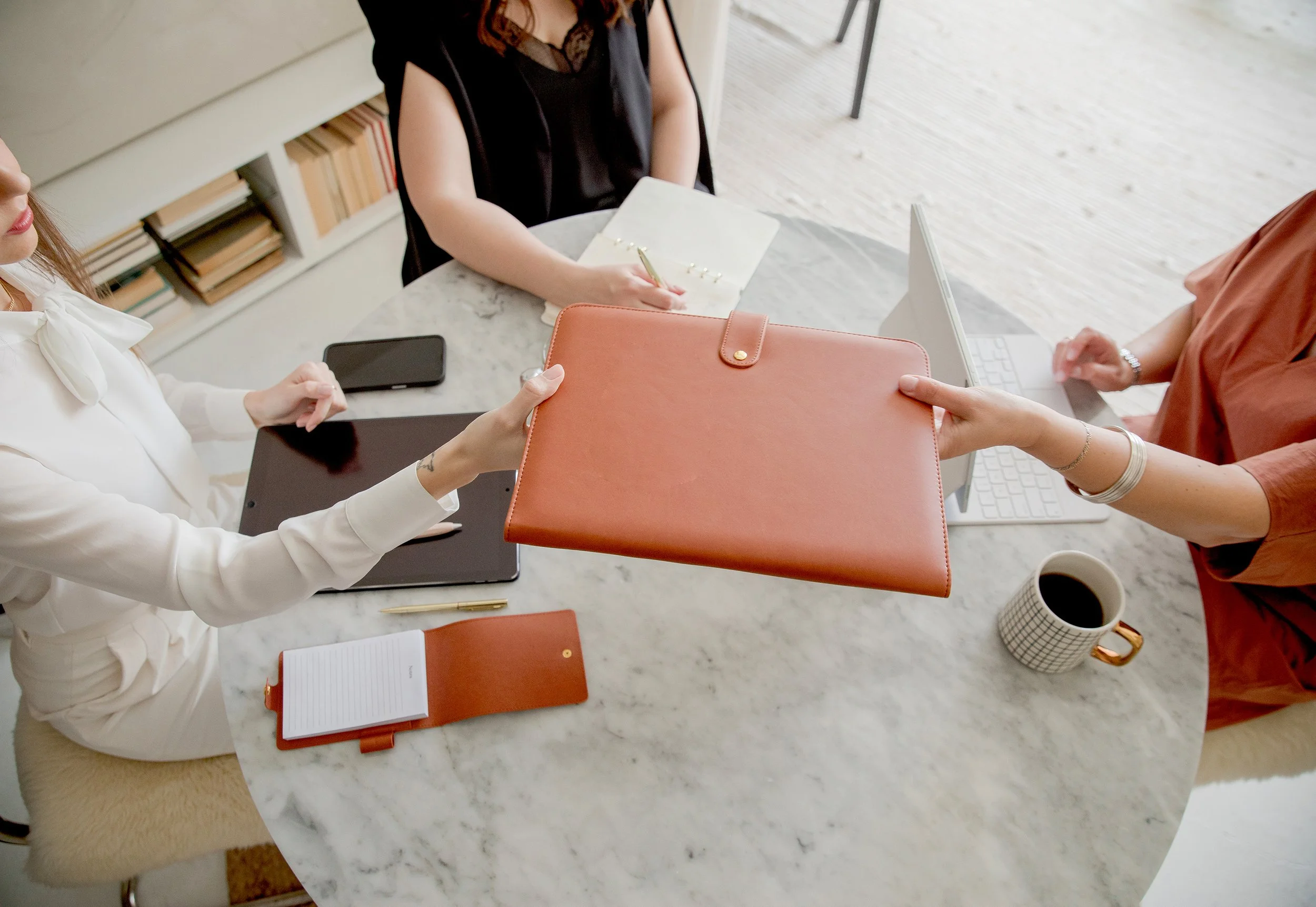 Three women sit around a round marble table in a modern office, exchanging a leather portfolio and discussing documents, with notebooks, a mug, and a mobile phone on the table.