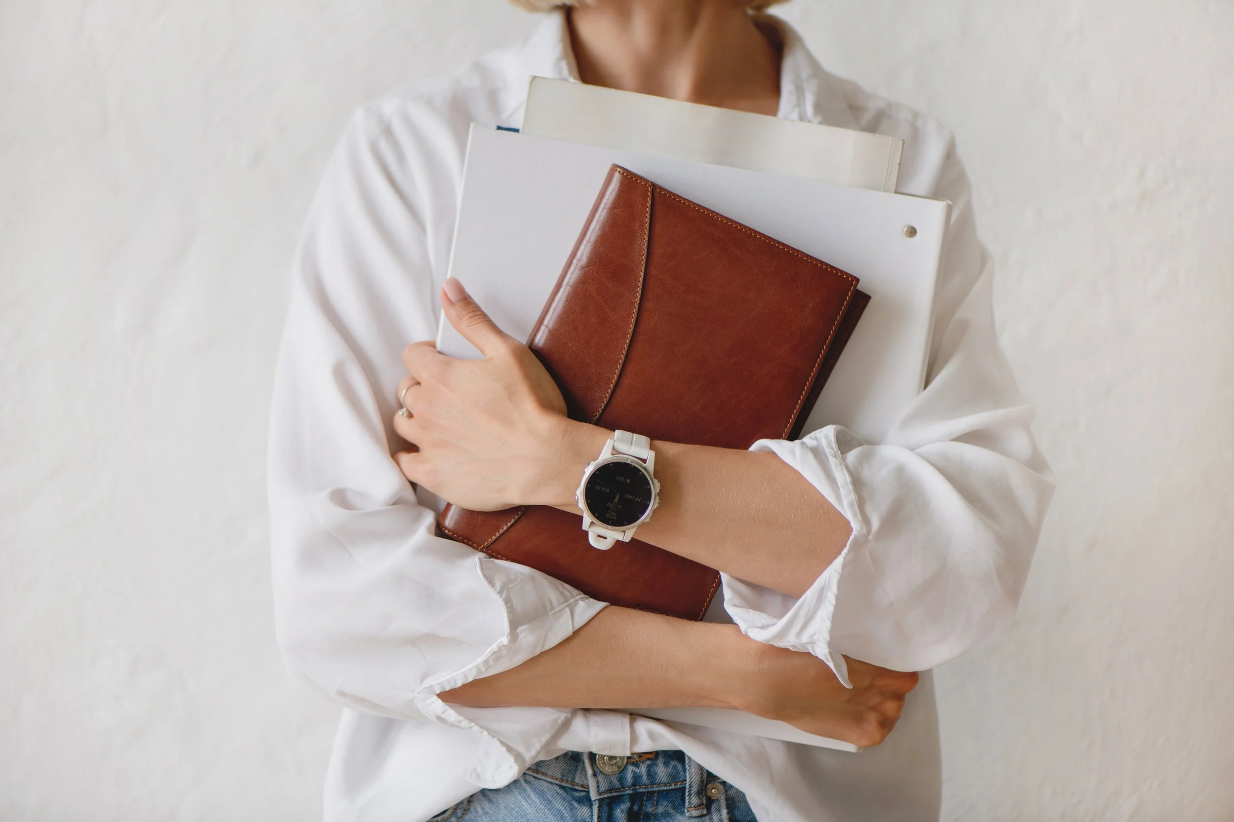 Person holding a brown leather notebook and three white books, wearing a white button-up shirt, a watch, and rings, standing against a plain white wall.