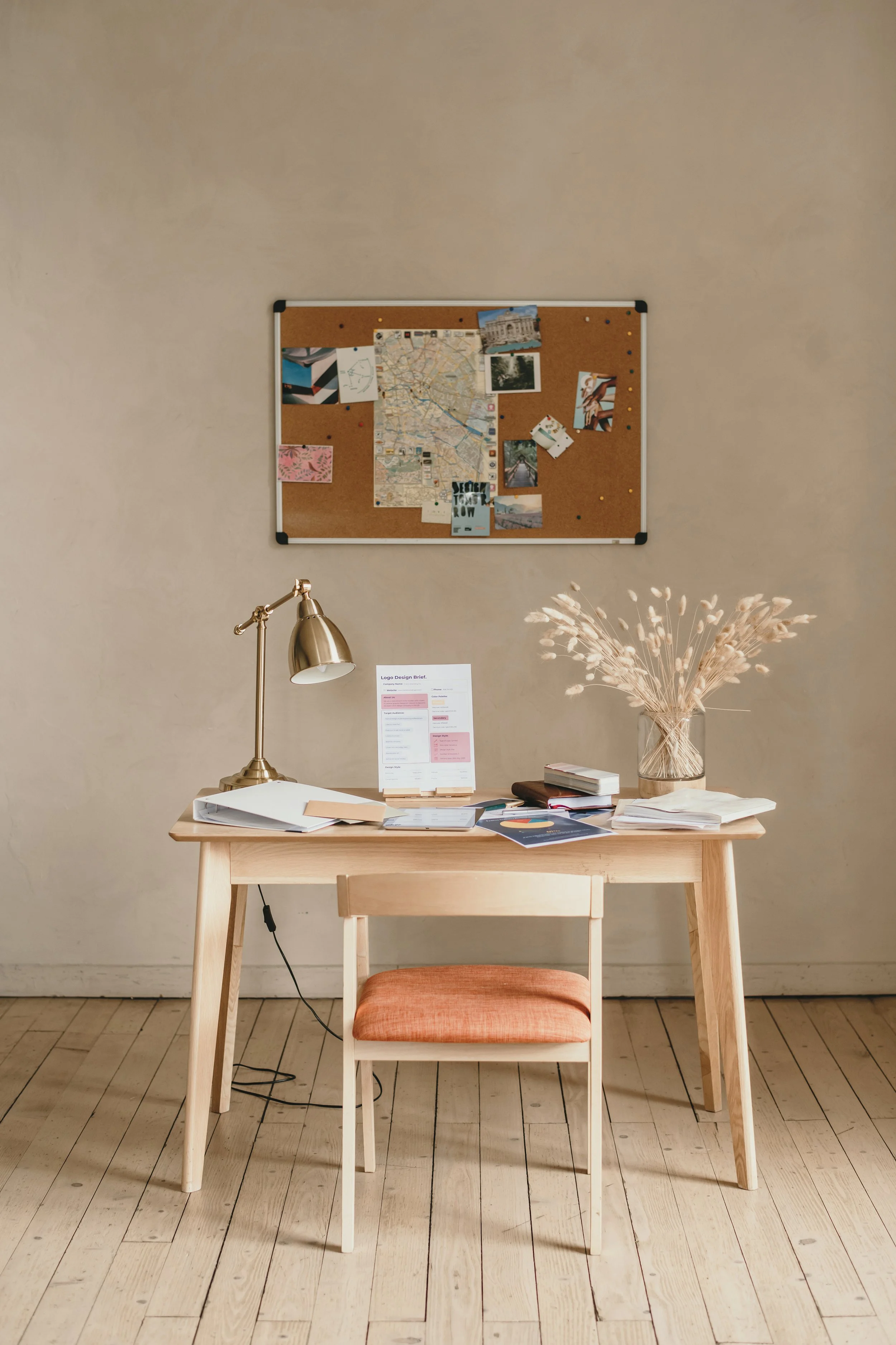 A workspace with a wooden desk, a matching wooden chair with an orange cushion, a white desk lamp, a glass vase with dried plants, and various papers and documents on the desk. A corkboard with photos and a map is mounted on the beige wall above the desk.