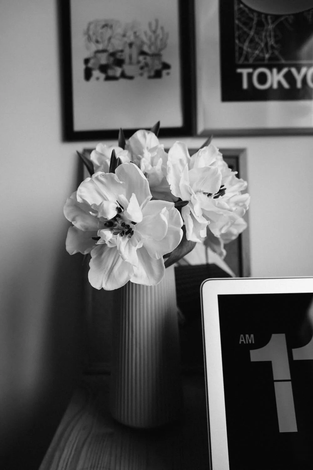 Black and white photo of a vase with flowers on a wooden surface, with framed artwork on the wall behind and part of a digital clock or device screen visible in the foreground.