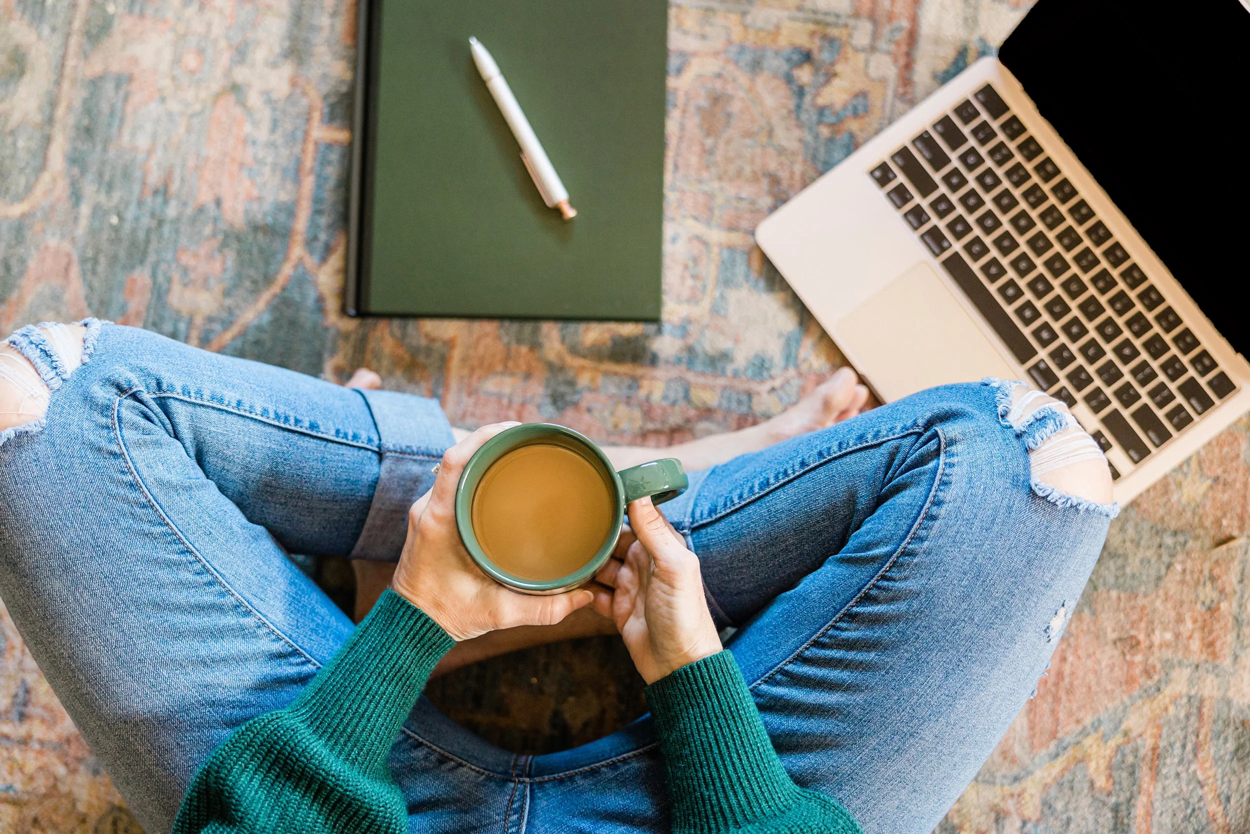 A person sitting on the floor with legs crossed, wearing ripped blue jeans and a green sweater, holding a green mug of coffee. In front of them on the floor is an open laptop, a closed dark green notebook, and a white pen. The floor is covered with a patterned area rug.