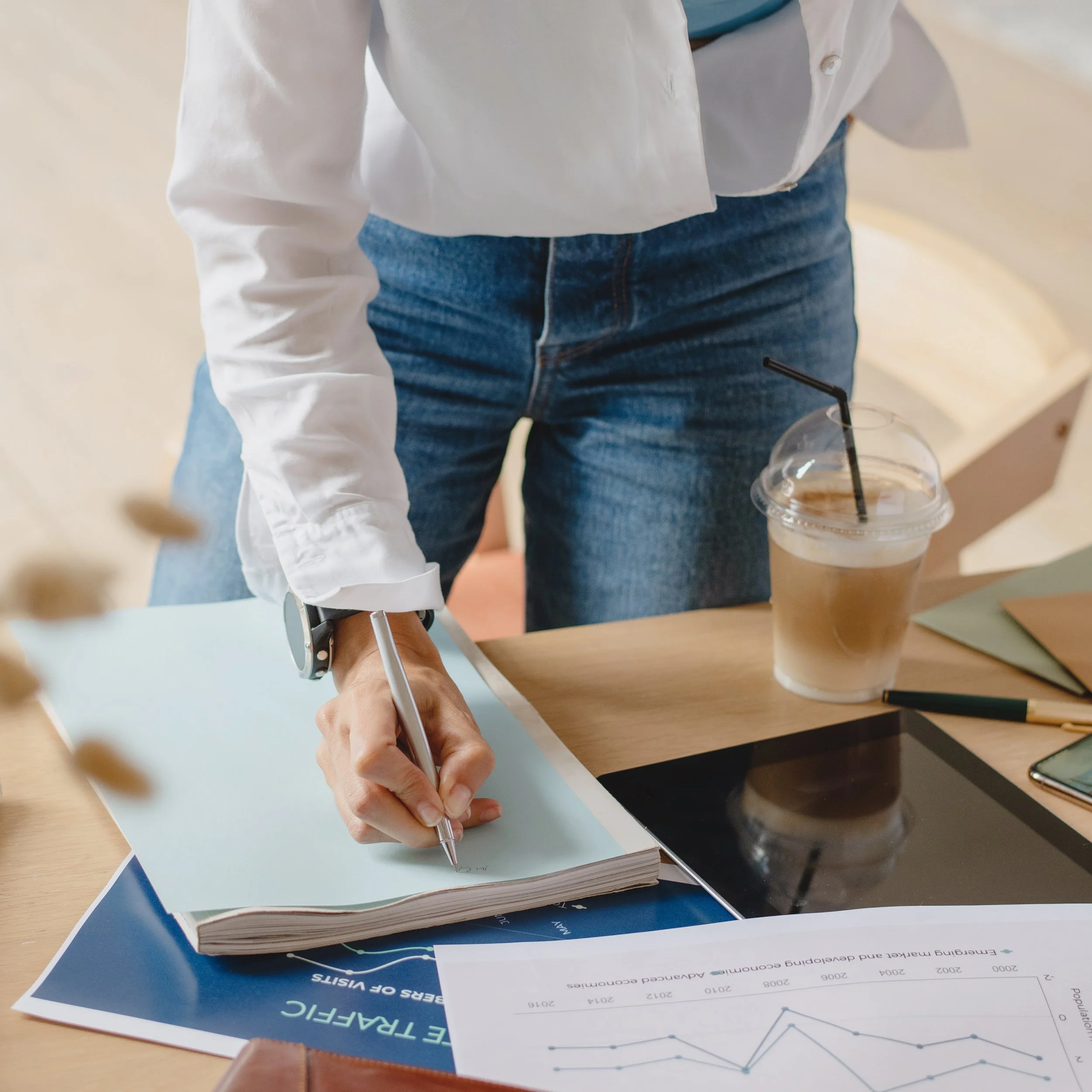 Person in white shirt and jeans writing notes on paper at a desk, with reports and documents nearby, and a beverage with a straw in the foreground.