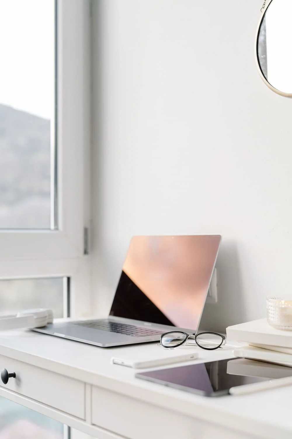 Modern workspace with a laptop, glasses, and tablet on a white desk near a window in a bright room.