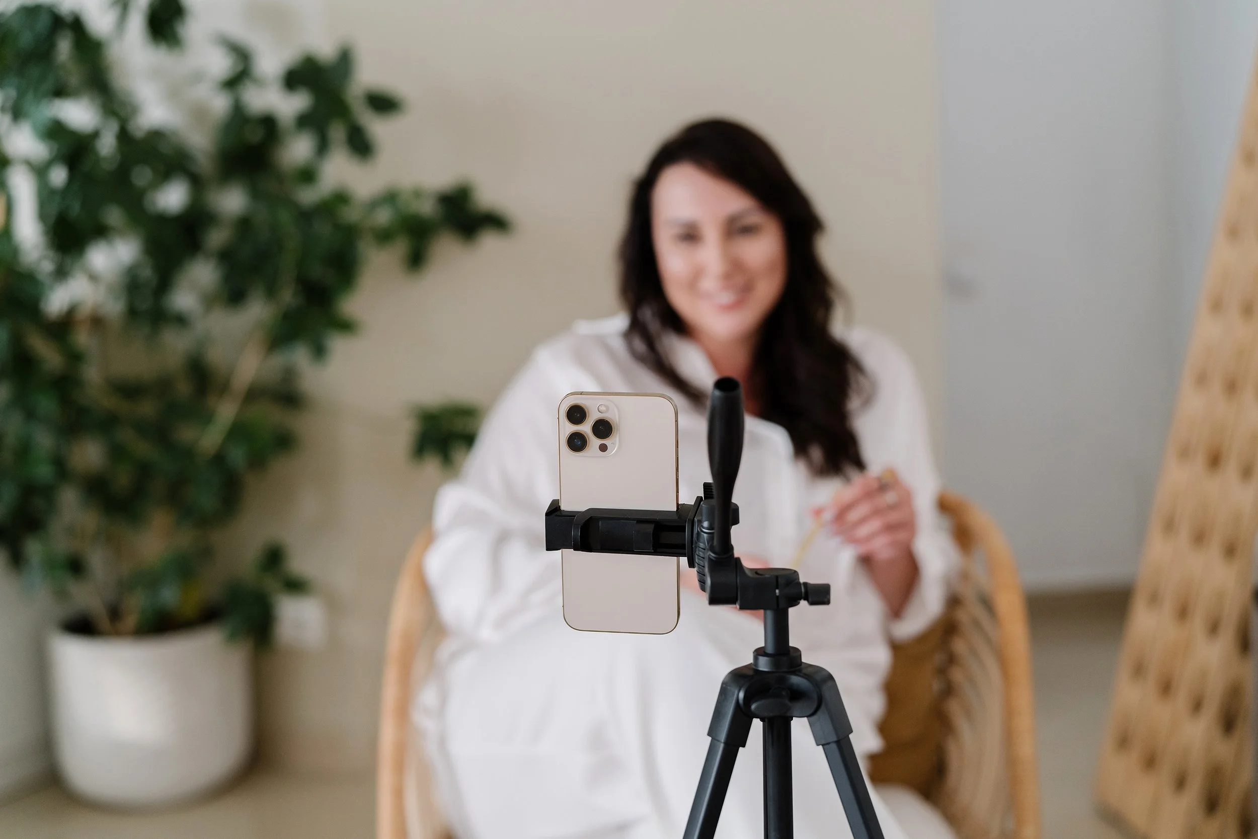 A woman with long dark hair sitting in a room, smiling, with a smartphone mounted on a tripod in front of her, taking a selfie or recording a video.