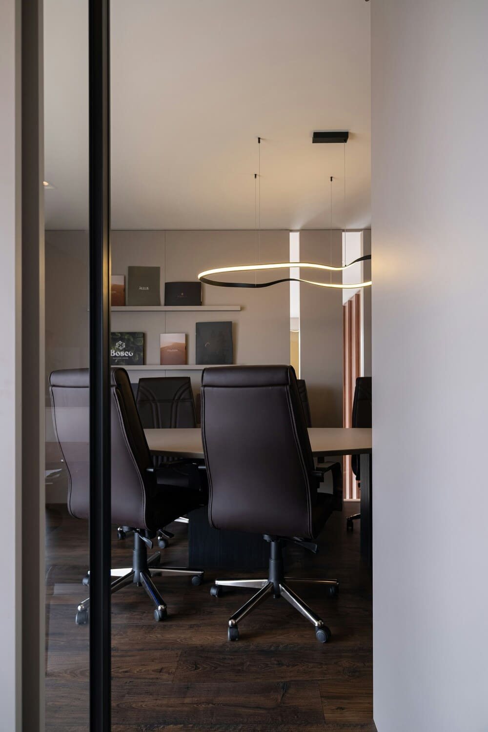 Modern conference room with black leather office chairs around a beige table, books on a white shelf, and a unique LED light fixture on the ceiling.