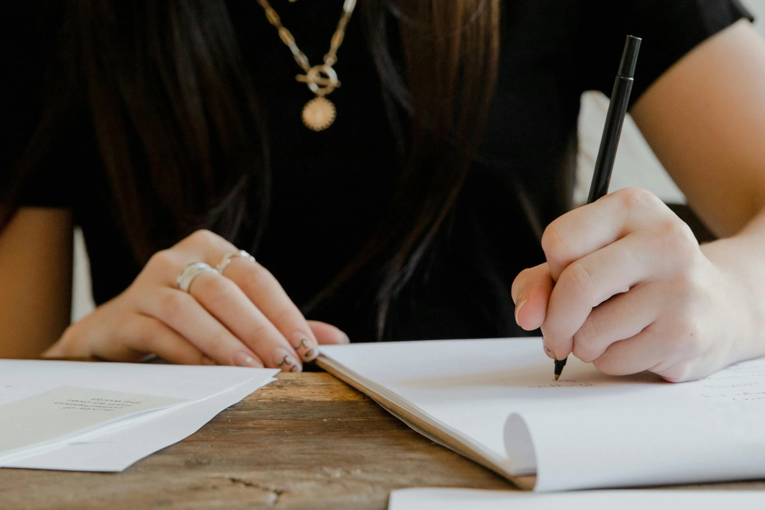 A person writing in a notebook with a black pen, wearing rings and a gold necklace, at a wooden table.