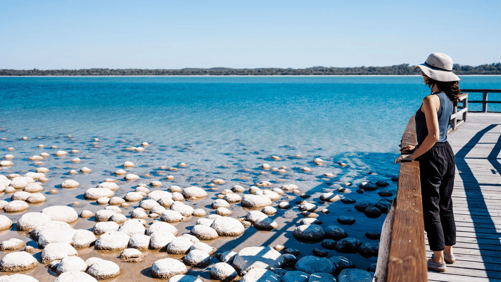 A woman standing on a wooden pier looking at the calm blue water and distant shoreline, wearing a hat and black clothing on a sunny day.