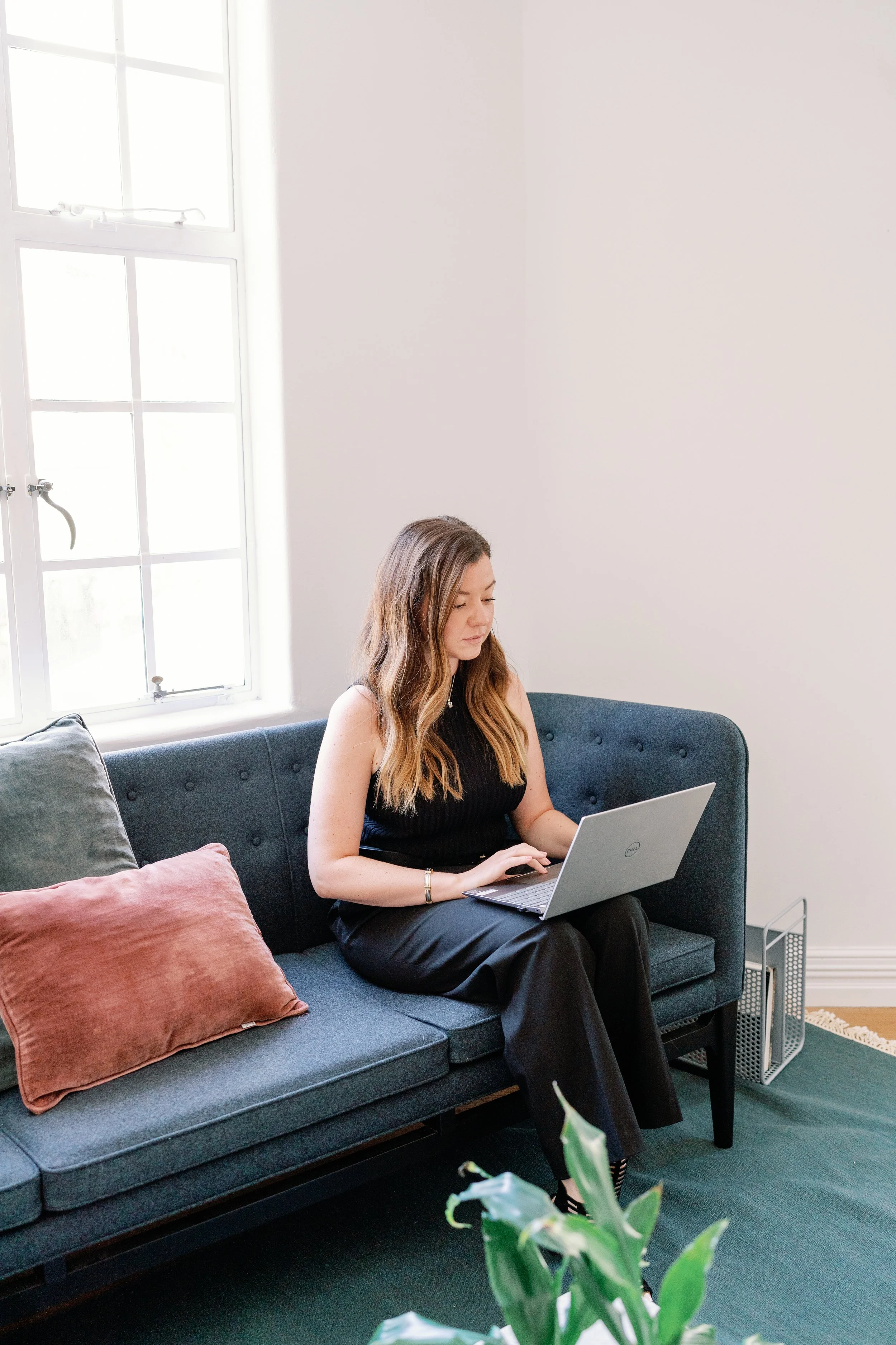 A woman with long wavy hair sitting on a dark gray sofa, working on a silver laptop in a bright, minimalist room with white walls and a window with divided panes.