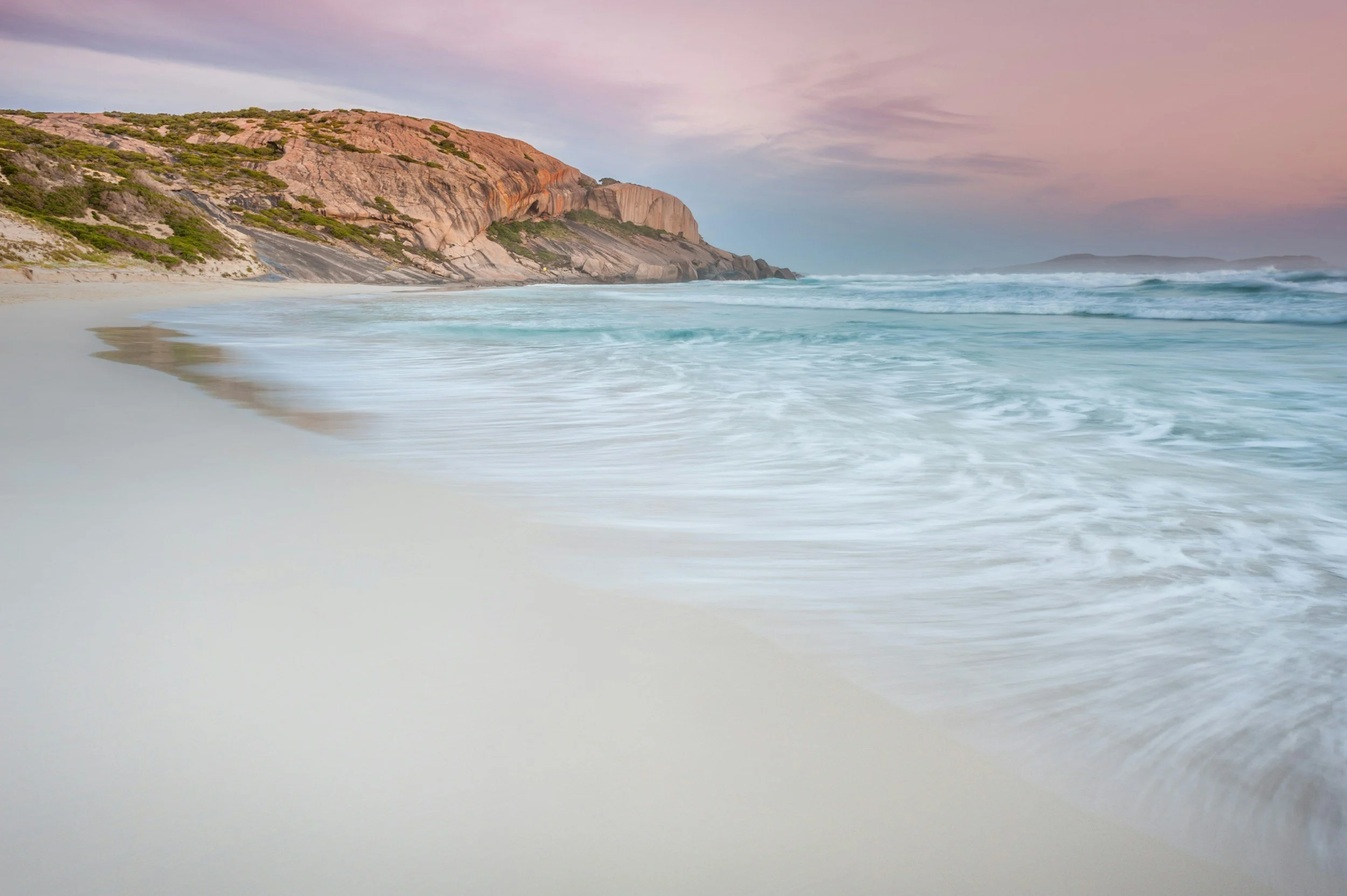 A scenic view of a beach with white sand, light blue waves, rocky cliffs with greenery, and a pinkish-purple sky at sunset.