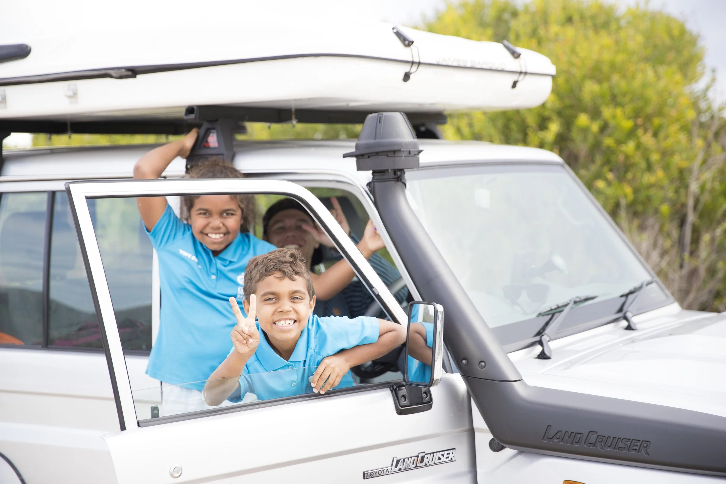 Kids riding in a white Toyota Land Cruiser with open windows, smiling and making peace signs, outdoors with green trees in the background.