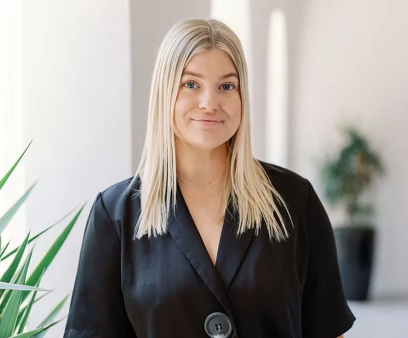 A young woman with long blonde hair, wearing a black blazer, standing indoors with a slight smile, in front of a blurred background with greenery.