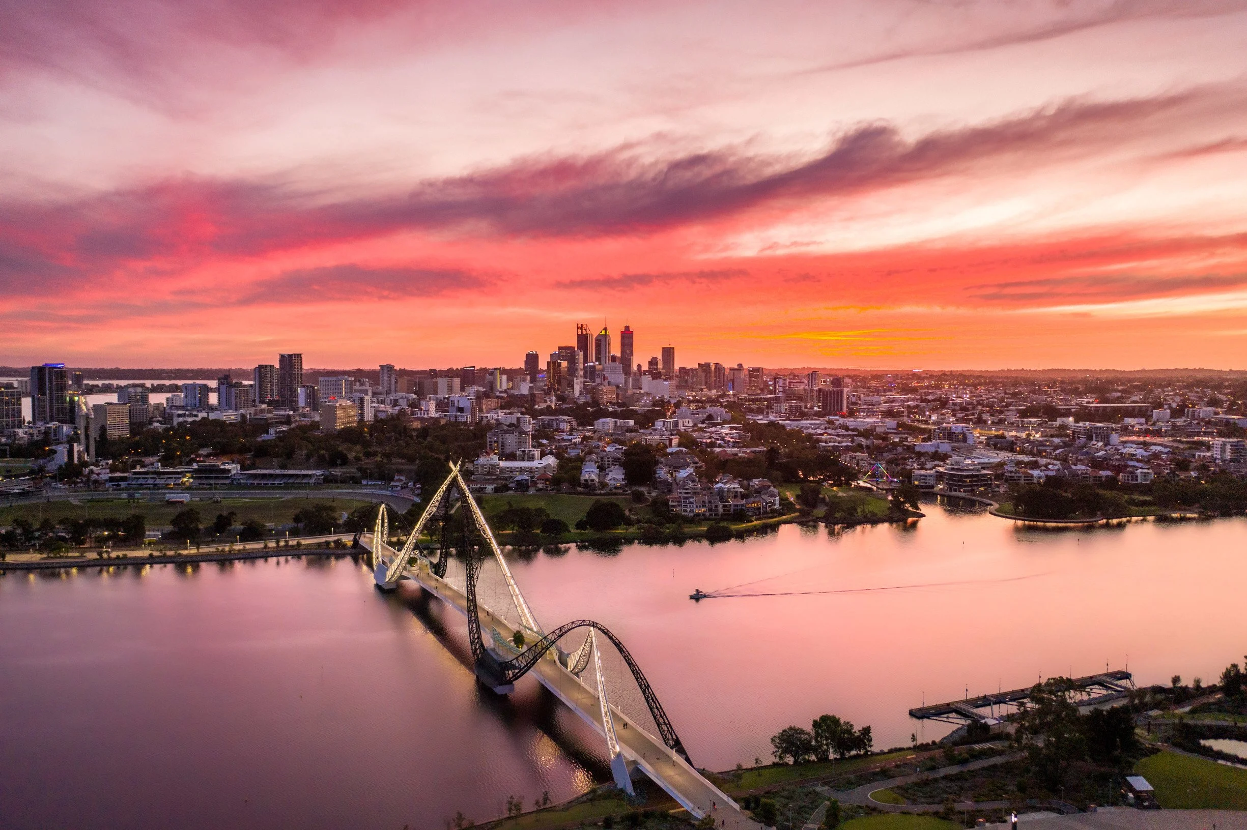 A city skyline at sunset with pink and purple clouds, a river in the foreground, and a bridge over the river.