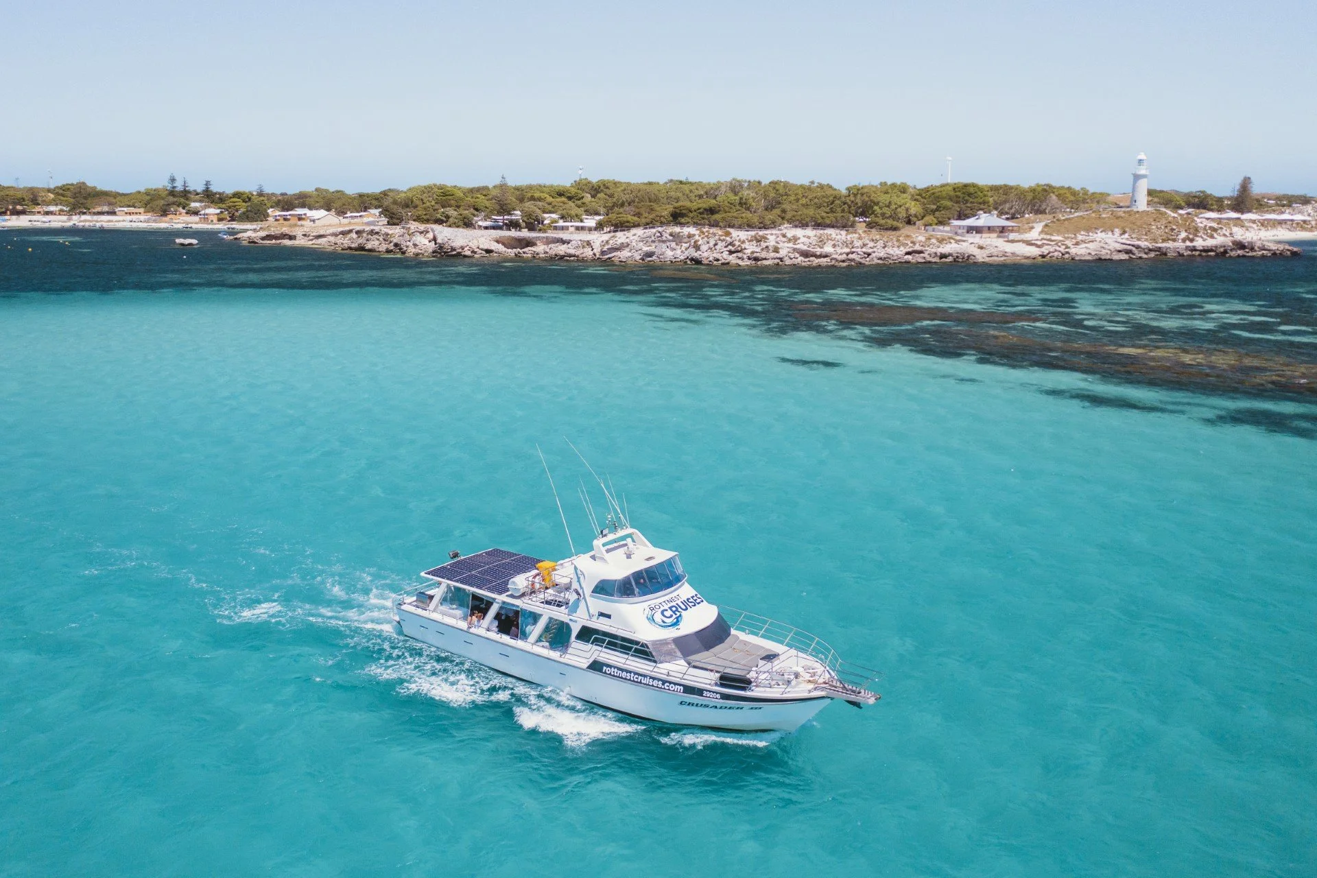 A white motor yacht moving through turquoise water near a rocky shoreline with green trees and a lighthouse in the background during daytime.
