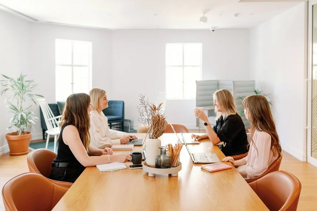 Five women sitting around a large wooden conference table in a bright, modern office, engaged in a discussion or meeting.