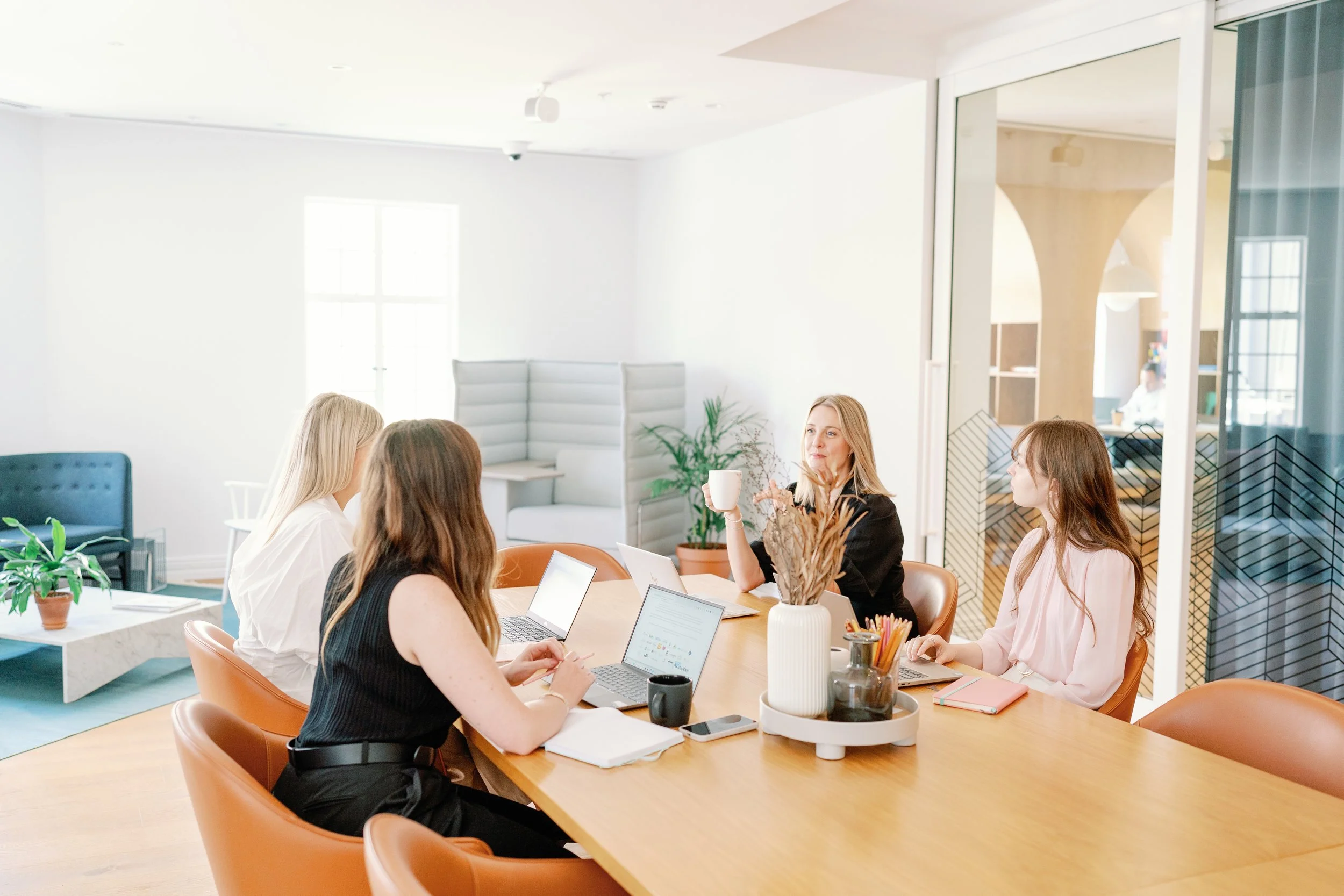 A group of women sitting around a conference table in a bright, modern office, engaged in discussion with laptops and notebooks, with plants and a glass partition in the background.