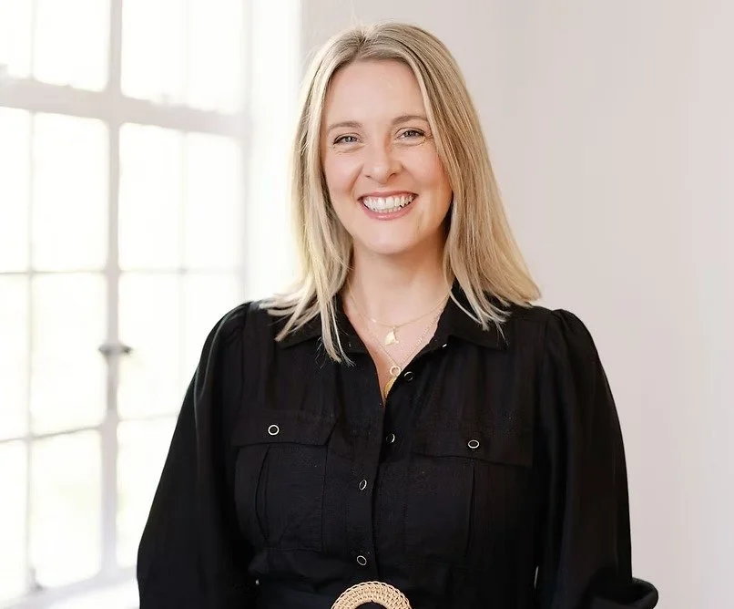 A woman with blonde hair smiling, wearing a black blouse and jewelry, standing in front of a large window with white frames.