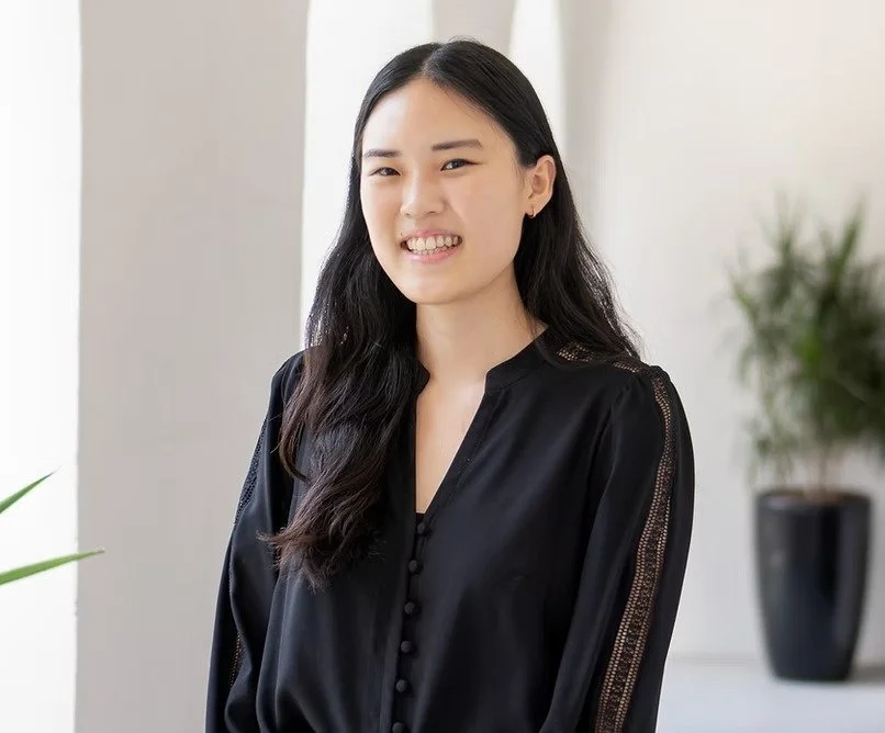 Young woman with long black hair wearing a black blouse, smiling in a bright indoor setting with potted plant in the background.