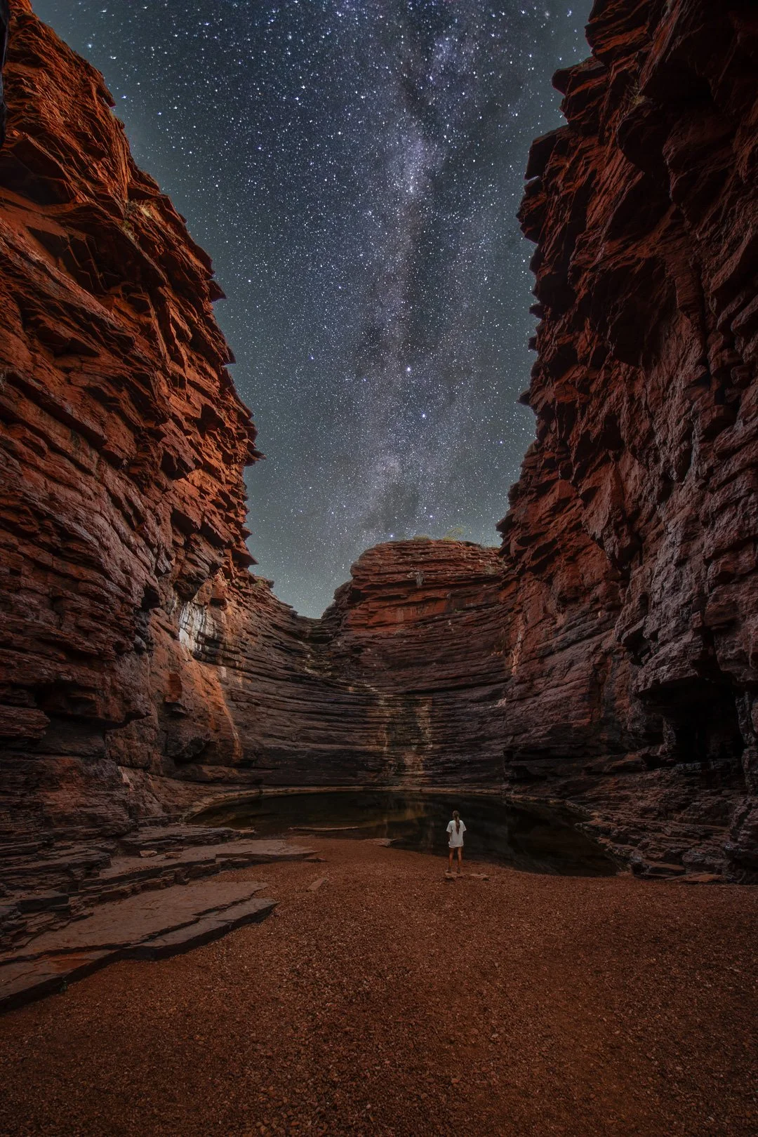 A person standing at the bottom of a crevice canyon with red rock walls, looking up at a starry night sky with the Milky Way galaxy visible.