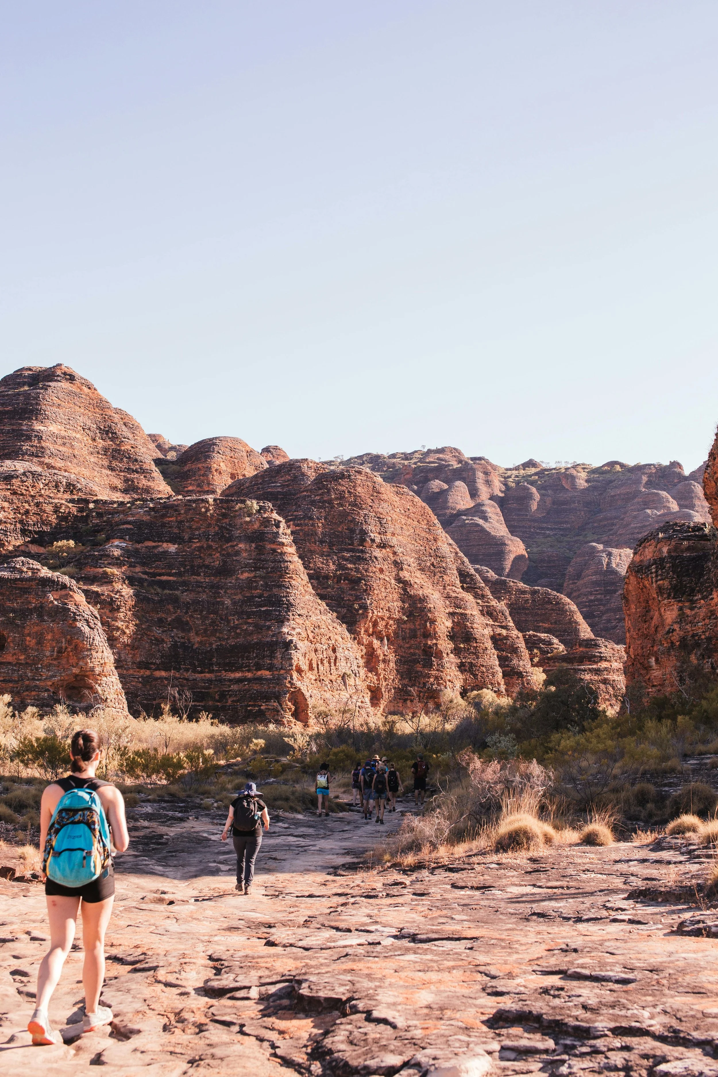 Hikers walking through a desert landscape with large reddish rock formations under a clear sky.