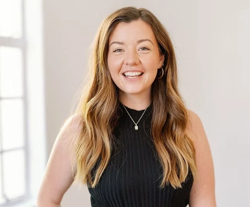 A smiling young woman with long wavy brown hair, wearing a black sleeveless top, silver necklace, and hoop earrings, standing indoors near a window.